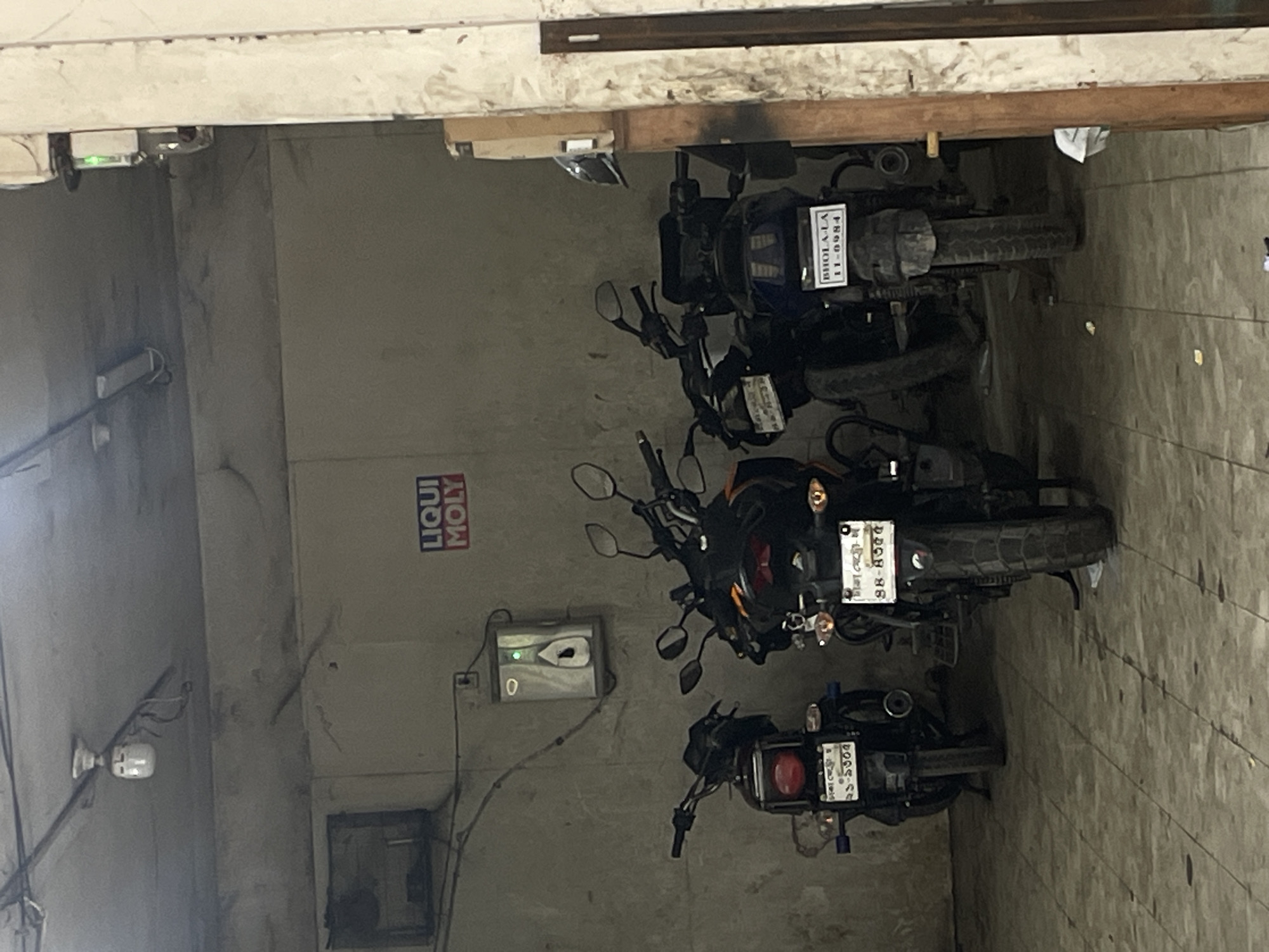 The image shows an indoor parking area, possibly a garage, with three motorcycles parked in a row. On the wall is a "LIQUI MOLY" logo and an electrical device with a green light. One motorcycle has a license plate that reads "ঢাকা যেতো ২১-১৩০৫", another "ঢাকা মেট্রো-অ ৪৪-৪৩৫৫", and the third "BHOLA LA 11-0984". These license plate inscriptions indicate the location is likely in Dhaka, Bangladesh. The environment appears to be utilitarian and possibly commercial, with a painted wall, exposed wiring, and tiled floor.