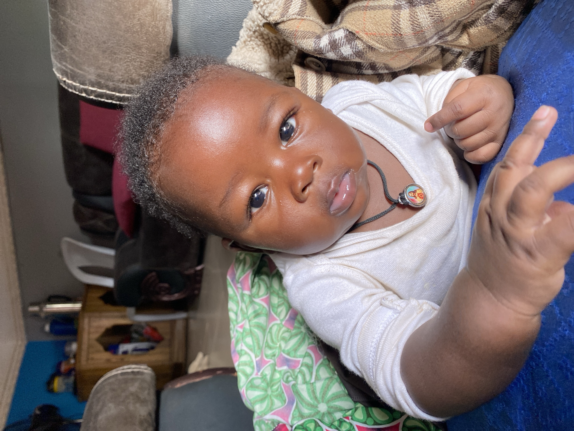 An eye-level, close-up photograph captures an infant lying down in an indoor setting in Vom, Nigeria. The baby, with dark skin, short curly black hair, and expressive dark eyes, looks directly at the camera with a neutral, slightly curious expression and slightly parted lips. The child wears a plain white long-sleeved bodysuit and a thin dark cord necklace with a small, round pendant. The pendant features a colorful, miniature image, possibly depicting two figures.
The baby is positioned amidst various textiles. To the upper left, a brown and beige plaid blanket or fabric is visible, while a vibrant green and pink patterned fabric lies beneath the baby on the lower left. To the baby's right, a textured blue fabric or cushion provides support. The background hints at a cluttered but lived-in environment, with glimpses of dark brown furniture, a wooden surface, and blurry household items on a blue mat. The lighting is soft and bright, suggesting it is daytime. The image conveys a sense of candid intimacy, focusing on the baby's features and calm demeanor. No distinct text is visible in the frame.