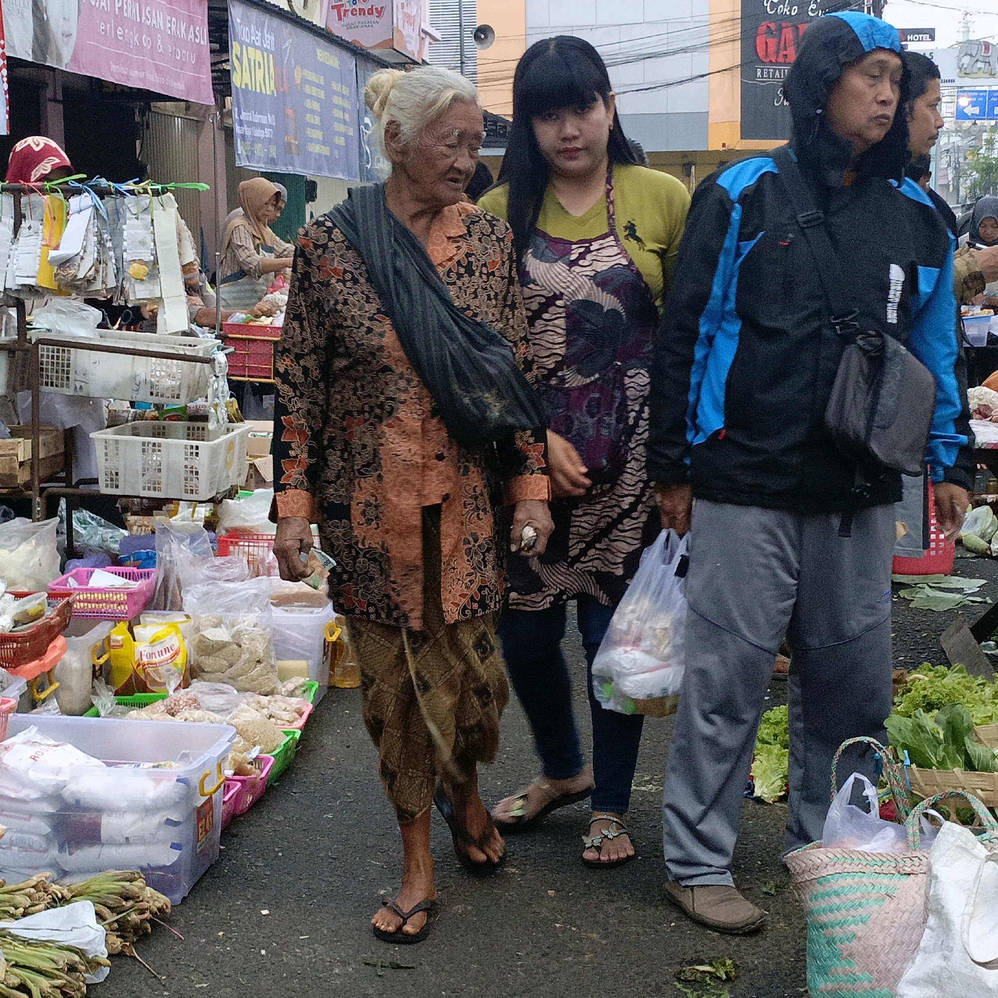 The image depicts a street market scene in Salatiga, Indonesia. In the foreground, an elderly woman with white hair and a traditional patterned orange and black blouse is visible from the chest down, wearing a dark shawl draped over her shoulder and a brown patterned skirt. She is standing next to a younger woman with long black hair, wearing a lime green top and a patterned apron. The younger woman is looking down and to the left. Further to the right, a person wearing a blue and black hooded jacket with a black bag slung across their body stands with a plastic bag in their left hand. A second person, partially obscured, is standing behind them. The background reveals market stalls laden with various goods, including plastic containers, baskets filled with food items such as leafy greens, packaged snacks, and bottles of oil. Signs in Indonesian are visible above the stalls. The ground is paved and appears wet, suggesting recent rain. The overall atmosphere is that of a bustling local market.