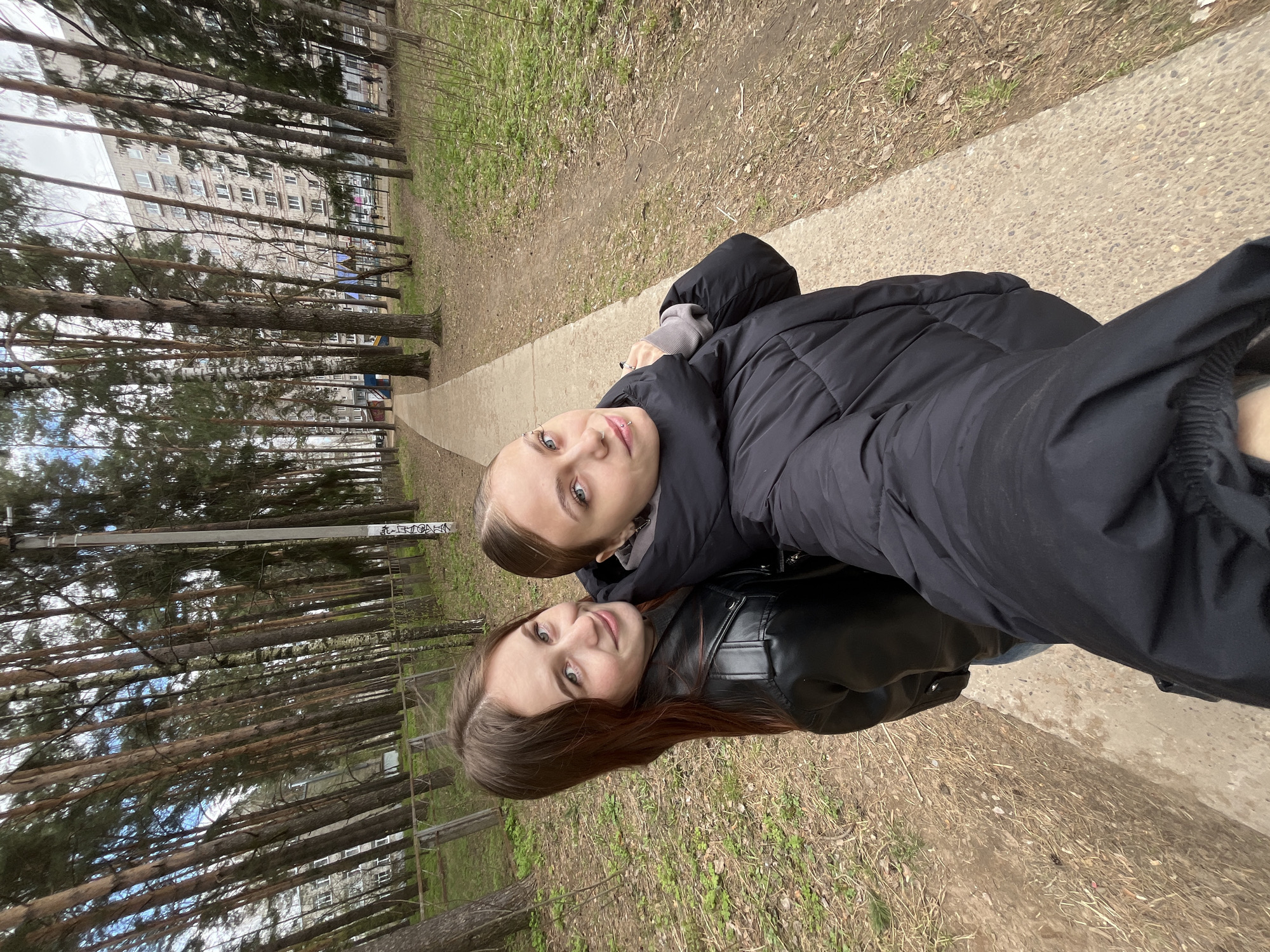 Two women are standing on a concrete path in a park-like area. The woman on the left is wearing a black leather jacket and has brown hair. The woman on the right is wearing a black puffer jacket and has blonde hair. Trees are visible in the background, as is a building. The photograph appears to have been taken in Chaikovsky, Russia.