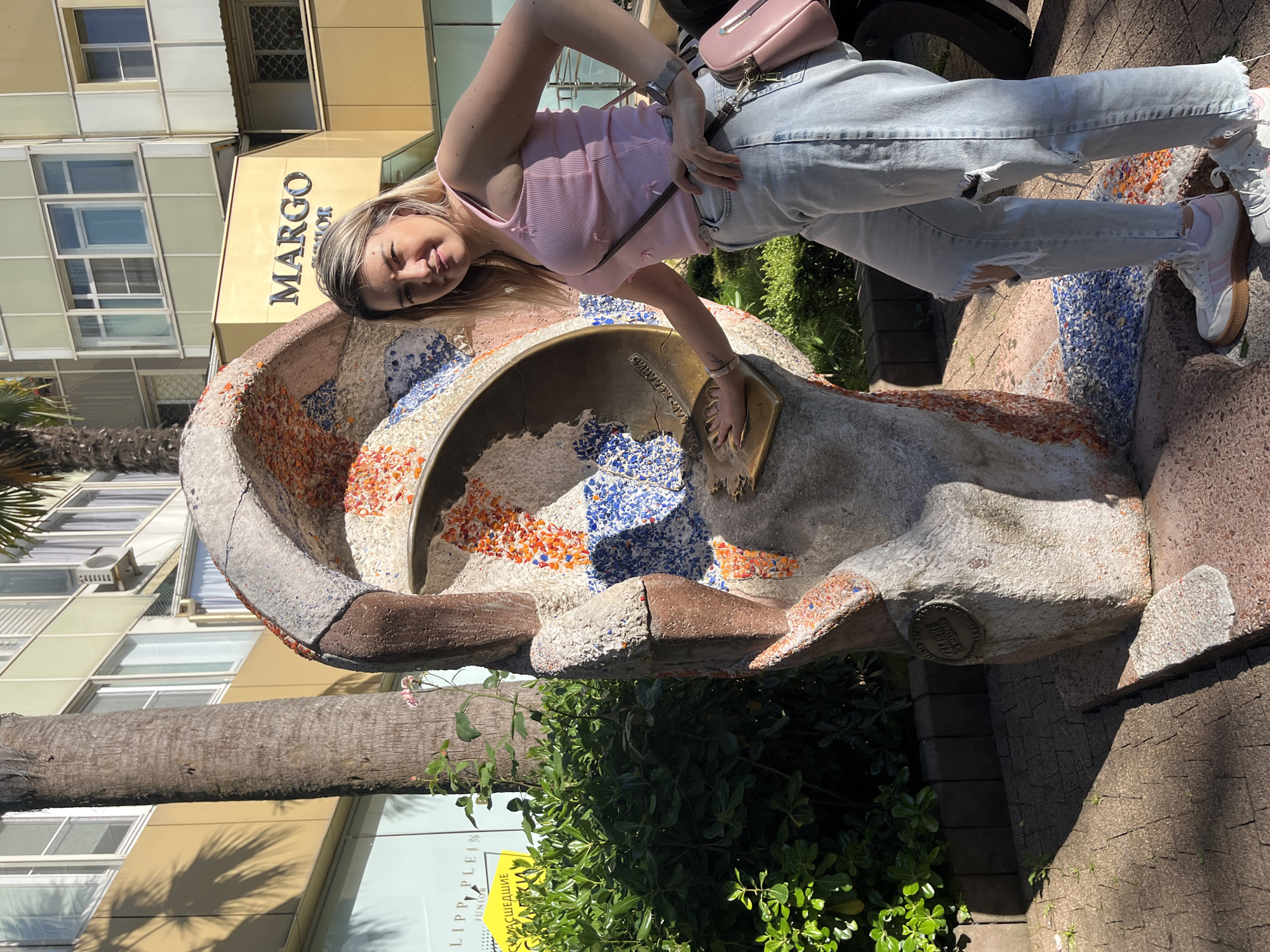 In Sochi, Russia, a young Caucasian woman with blonde hair and light-colored eyes poses next to a large, sculpted ear artwork adorned with colorful mosaics. She is wearing a pink ribbed tank top, light-wash ripped jeans, and white sneakers with pink accents. A small pink crossbody bag is slung over her shoulder. Her right hand rests on a bronze plaque embedded in the sculpture, which appears to be a handprint. Behind the sculpture, a palm tree trunk is visible, and further in the background, there are buildings with large windows and signage, including one that reads "MARGO" in large gold letters. Lush green bushes are situated to the left of the sculpture and the woman. The ground is paved with brick-like stones. The scene is outdoors, under bright sunlight, casting shadows.