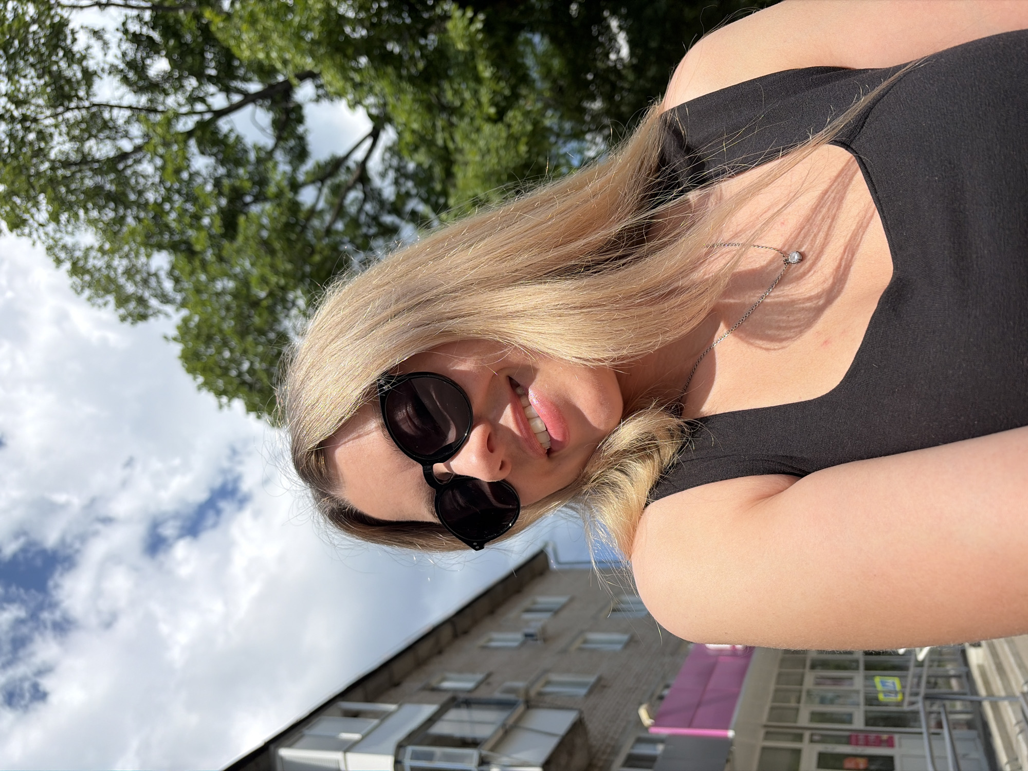 A young woman with blonde hair and sunglasses is taking a selfie outdoors in Chaikovsky, Russia. She is wearing a black tank top and a silver necklace with a small pendant. Behind her, there is a multi-story building with balconies and windows, a green tree, and a bright blue sky with white clouds. The woman is smiling and looking at the camera. The lighting suggests it is daytime.