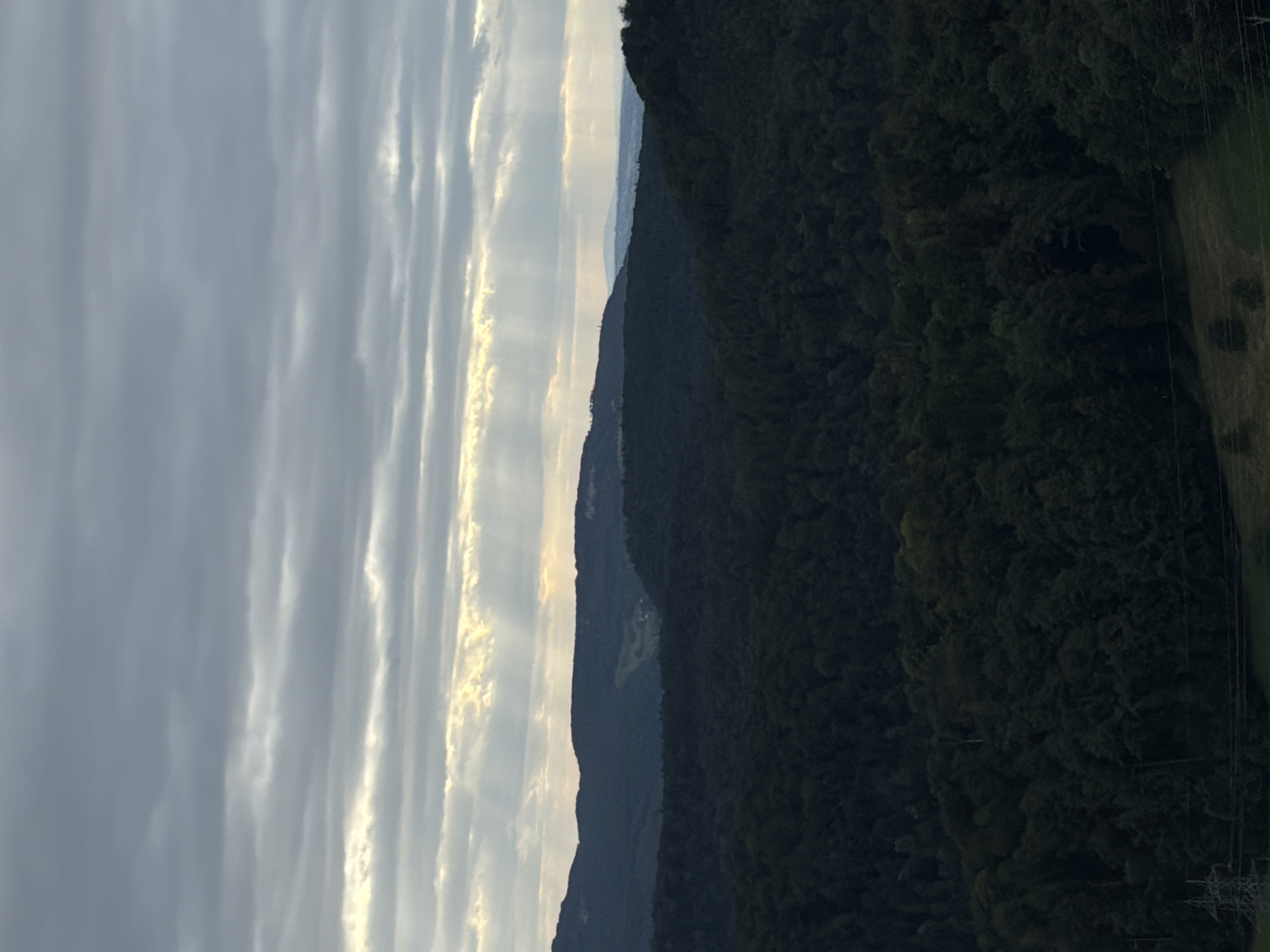 The image shows a landscape view from a high vantage point, likely in or around Sissach, Switzerland. The scene is dominated by rolling hills covered in dark green forests. In the distance, layers of hills recede into the background, with the furthest ones appearing hazy and a lighter blue, suggesting a broad valley or plain. The sky is overcast with dramatic clouds, through which shafts of sunlight are breaking, creating visible light rays. The light rays are most prominent in the upper portion of the image, illuminating patches of the sky and the upper slopes of the hills. In the foreground, a power pylon and some electrical lines are visible on the left side, indicating human infrastructure present in the otherwise natural landscape. The overall atmosphere is moody and somewhat dramatic due to the interplay of dark shadows and bright light. There are no people or animals visible in the frame.
