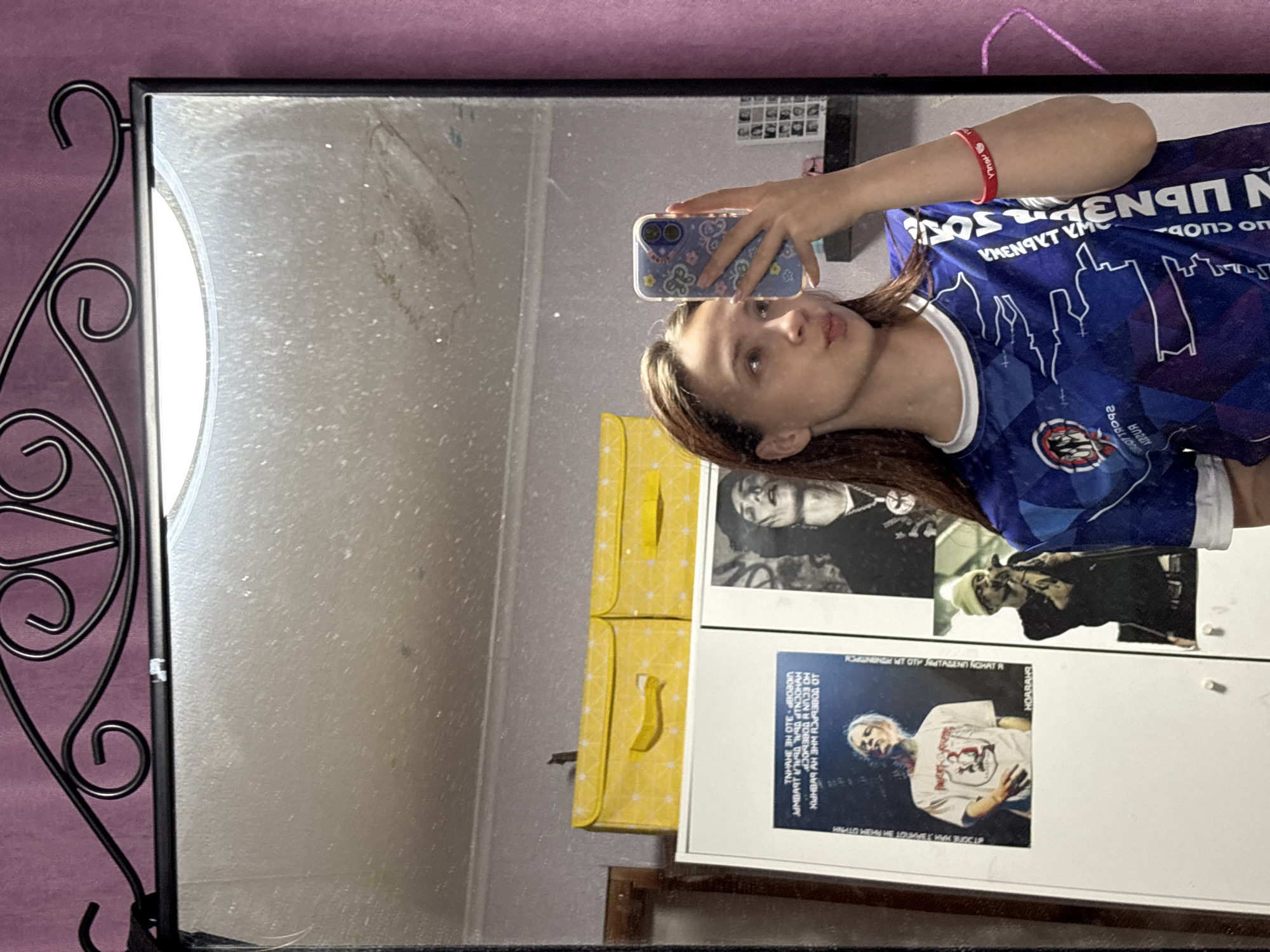 A mirror selfie shows a young woman in a blue soccer jersey with a red wristband. The reflection also shows a part of the room's decor. In the background, there is a yellow storage unit. Two black-and-white photographs are mounted on a white wall or cabinet, and the text near the bottom appears to be written in Russian. The surrounding environment includes a light-colored wall and a pink-purple background. The mirror has a decorative black frame with curlicue elements.