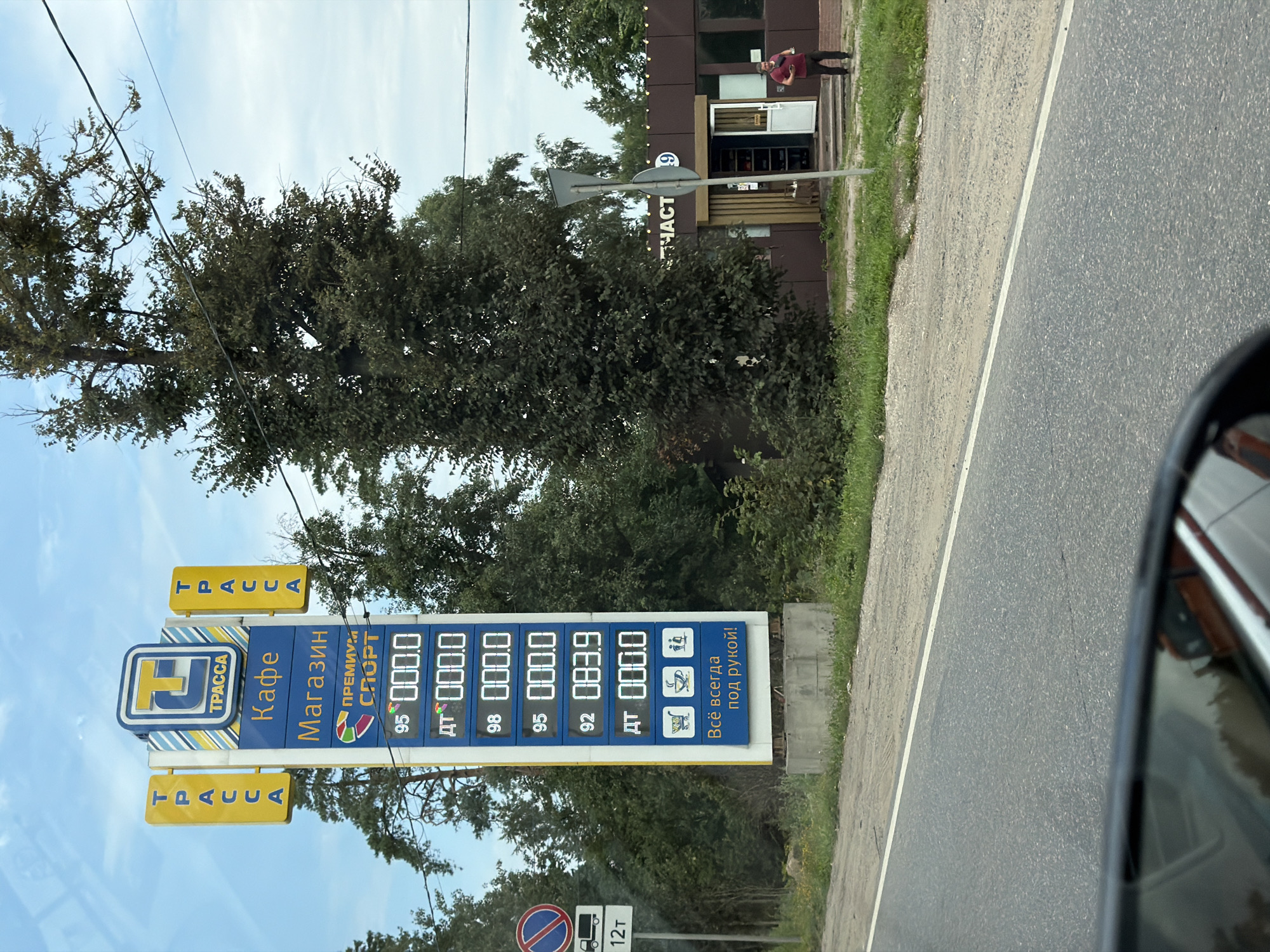 A vertical, rectangular sign on a concrete base displays fuel prices and services. The sign is predominantly blue and yellow, with a large blue and yellow logo at the top that reads "ТРАССА." Below the logo, it says "Кафе" (Cafe) and "Магазин" (Shop), followed by "ПРЕМИУМ" (Premium) and "СПОРТ" (Sport). The lower portion of the sign shows fuel prices displayed digitally. From top to bottom, the listed fuel types and prices are: 95 000.0, ДТ 000.0, 98 000.0, 95 000.0, 92 083.9, and ДТ 000.0. At the very bottom, there are icons depicting a shopping cart, a coffee cup, and people, with the text "Всё всегда под рукой!" (Everything is always at hand!). Two yellow vertical "ТРАССА" signs are attached to the left and right sides of the main sign. To the left of the main sign is a traffic sign with a red circle and a truck symbol, indicating a weight limit of 12 tons. A building with a brown facade and a light-colored awning is visible to the right of the sign. The building has an open doorway with a person in a red shirt standing outside. A street extends across the foreground, and a wooded area is visible behind the sign and building. The sky is partly cloudy. This scene is located in Radumlya, Russia, likely a roadside gas station.