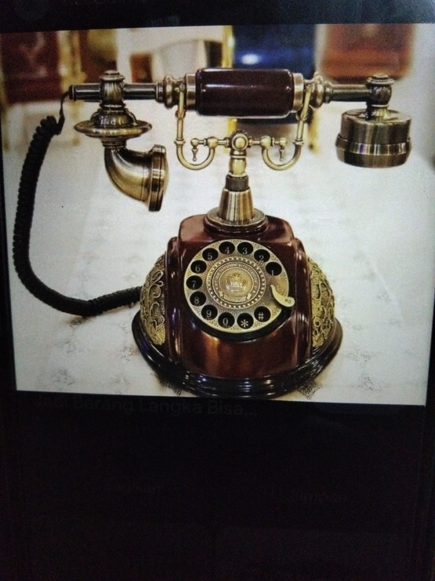 An antique rotary dial telephone with a dark brown body and brass accents sits on a white, textured surface. The telephone has a coiled black cord connecting the receiver to the base. The receiver itself is brass and has a dark brown handle. A circular dial with numbers zero through nine, as well as asterisk and hash symbols, is prominently featured on the front of the base. Decorative brass filigree adorns the sides of the base. The background is blurred but suggests an indoor setting with some architectural elements. The image has a watermark at the bottom that reads "Barang Langka Bisa". The location in the city of Kualaraja, Indonesia is not explicitly indicated by any visual cues in the image.