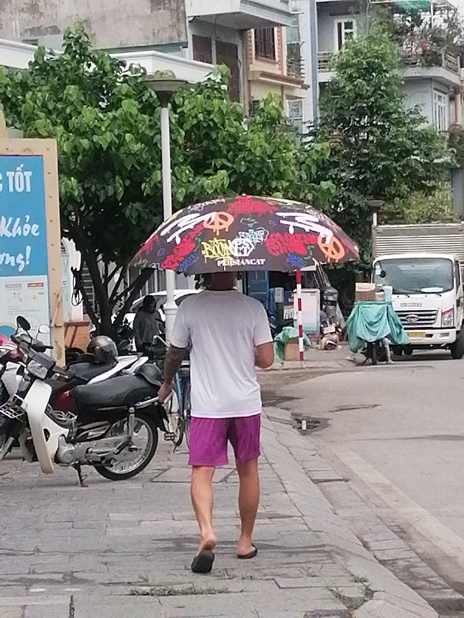 A man walks barefoot along a sidewalk on a street in Ha Tu Ward, Vietnam. He wears purple shorts and a white t-shirt, with a colorful umbrella balanced on his head. A motorcycle is parked nearby. In the background, there is a white truck parked on the street. The setting appears to be a residential street with trees and buildings.