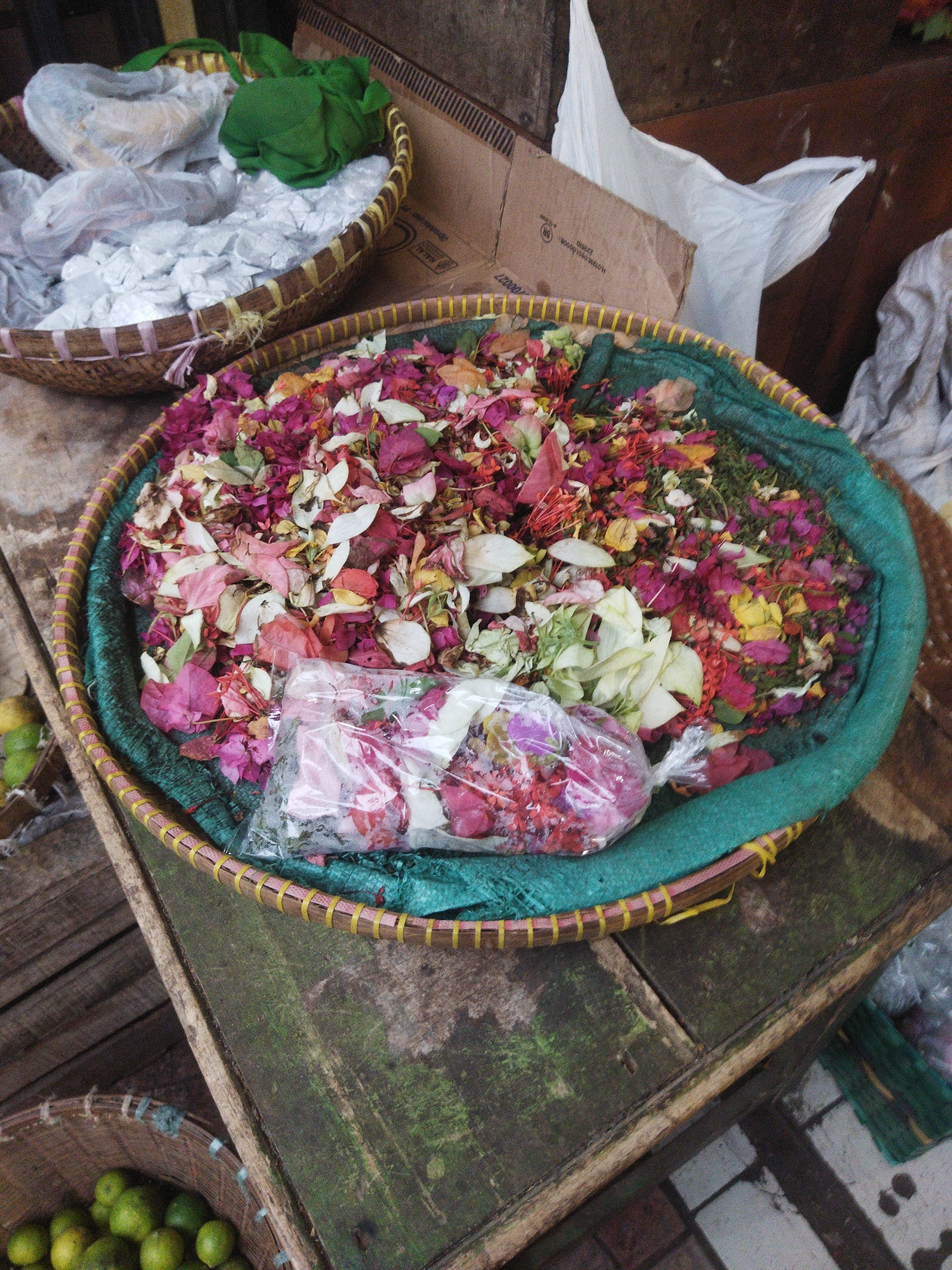 A high-angle, eye-level shot shows two woven baskets on a weathered wooden surface. The basket in the foreground, partially obscured by the edge of the frame, contains green limes. The basket to the upper left contains white items, possibly sugar packets, and a green fabric. The largest basket, in the center, is filled with dried flower petals in various shades of pink, red, yellow, and white, with a plastic-wrapped bundle of petals in the foreground. A brown cardboard box with writing is visible behind the baskets. This scene is likely from a market stall in the city of Banjar, Indonesia, given the common use of such baskets and the presence of fresh produce and dried flowers typical of Indonesian markets.