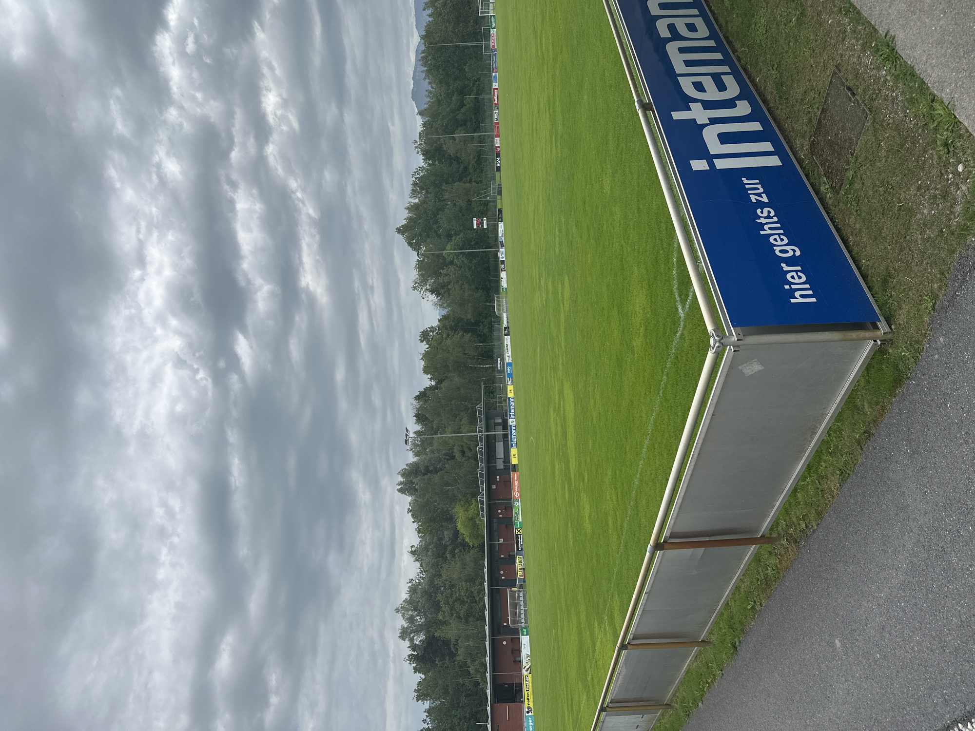 The image shows a football pitch in Lauterach, Austria, under a cloudy sky. The field is green with white markings, and a metal fence with a blue advertising banner runs along the side. The banner reads "hier gehts zur inteman". In the background, there is a row of trees and a building with a red facade. There are no people visible in the image.