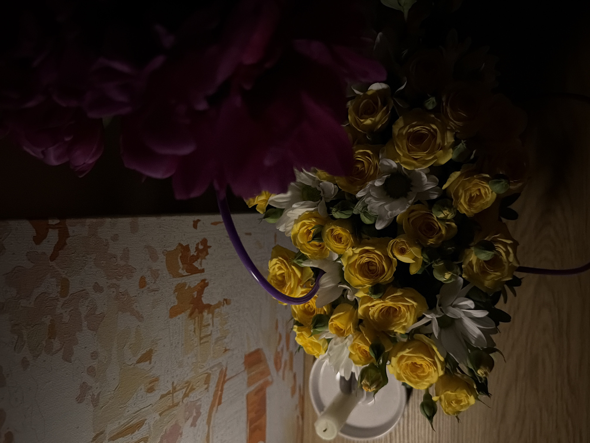 The image depicts a floral arrangement featuring yellow roses and white daisies, partially obscured by dark pink flowers in the upper left corner. The arrangement is placed on a white candle holder with a partially visible white candle. To the left of the flowers is a patterned surface, potentially a painted canvas or textured wall. A purple decorative element curves through the arrangement. The background consists of a dark wall on the right and possibly a continuation of the patterned surface on the left. The scene appears to be indoors, lit by dim, artificial light.