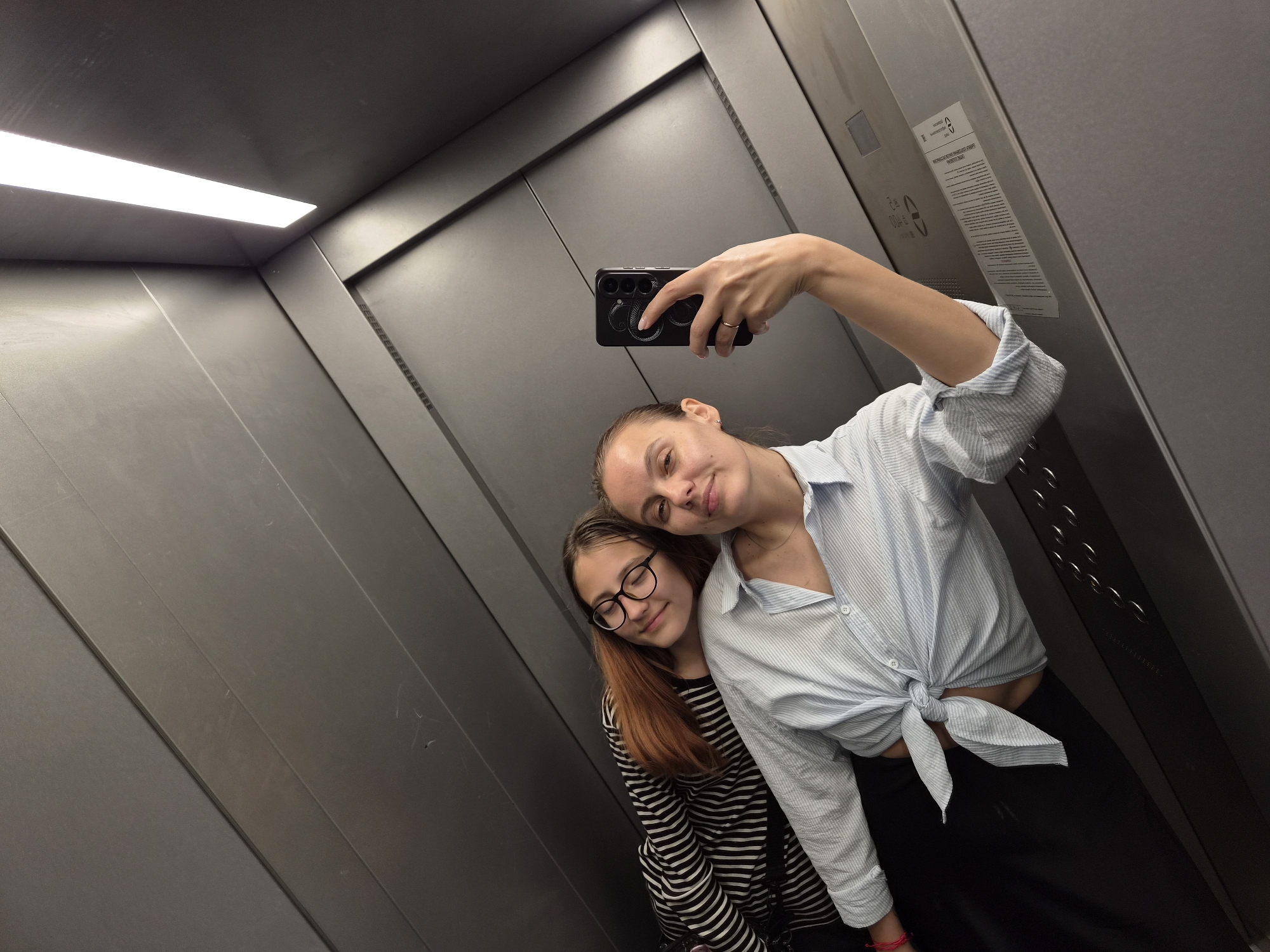 Two young women are taking a selfie in an elevator. The woman in the foreground is holding the phone, and the other woman is leaning on her shoulder. The elevator walls are metallic gray, with horizontal paneling and a bright fluorescent light at the top. The woman in the foreground is wearing a light blue, striped shirt tied at the waist, and black pants. The woman in the background is wearing a black and white striped shirt and glasses. The woman in the foreground has a ring on her right hand and a red bracelet on her left wrist.