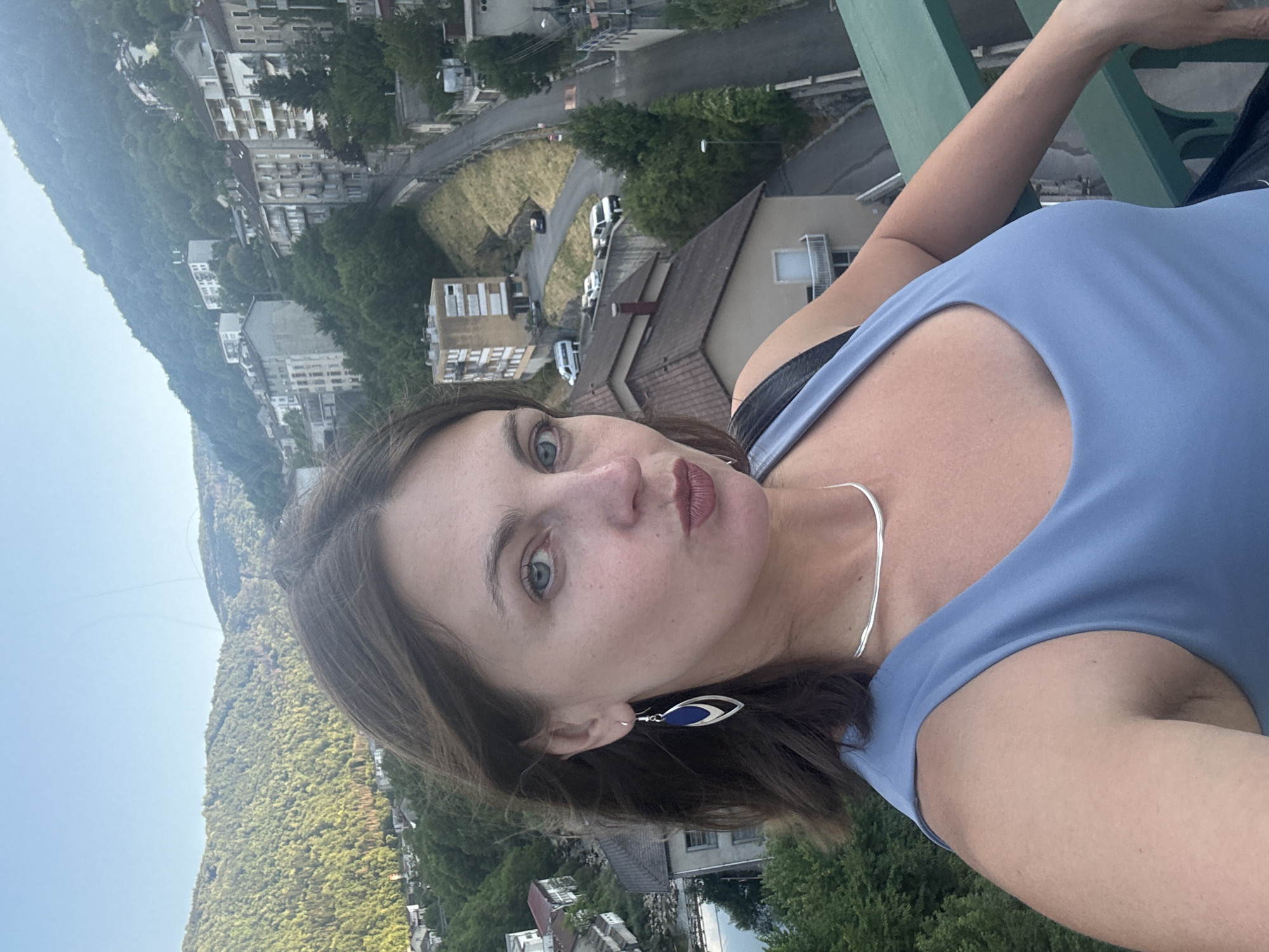 The image is a selfie taken outdoors, likely from an elevated position, overlooking a town nestled in a valley. The background features rolling, tree-covered hills and a cluster of buildings with a winding road. The architecture suggests a European town, consistent with Saint-Claude, France, known for its hilly terrain and urban development on slopes. In the foreground, a woman is captured from the chest up, facing the camera. She has medium-length brown hair, fair skin, and is wearing a blue sleeveless top and a silver necklace. Her gaze is directed at the camera, with her lips pursed. She is also wearing a pair of distinctive blue and silver dangling earrings. The overall lighting suggests it is either late afternoon or early evening.