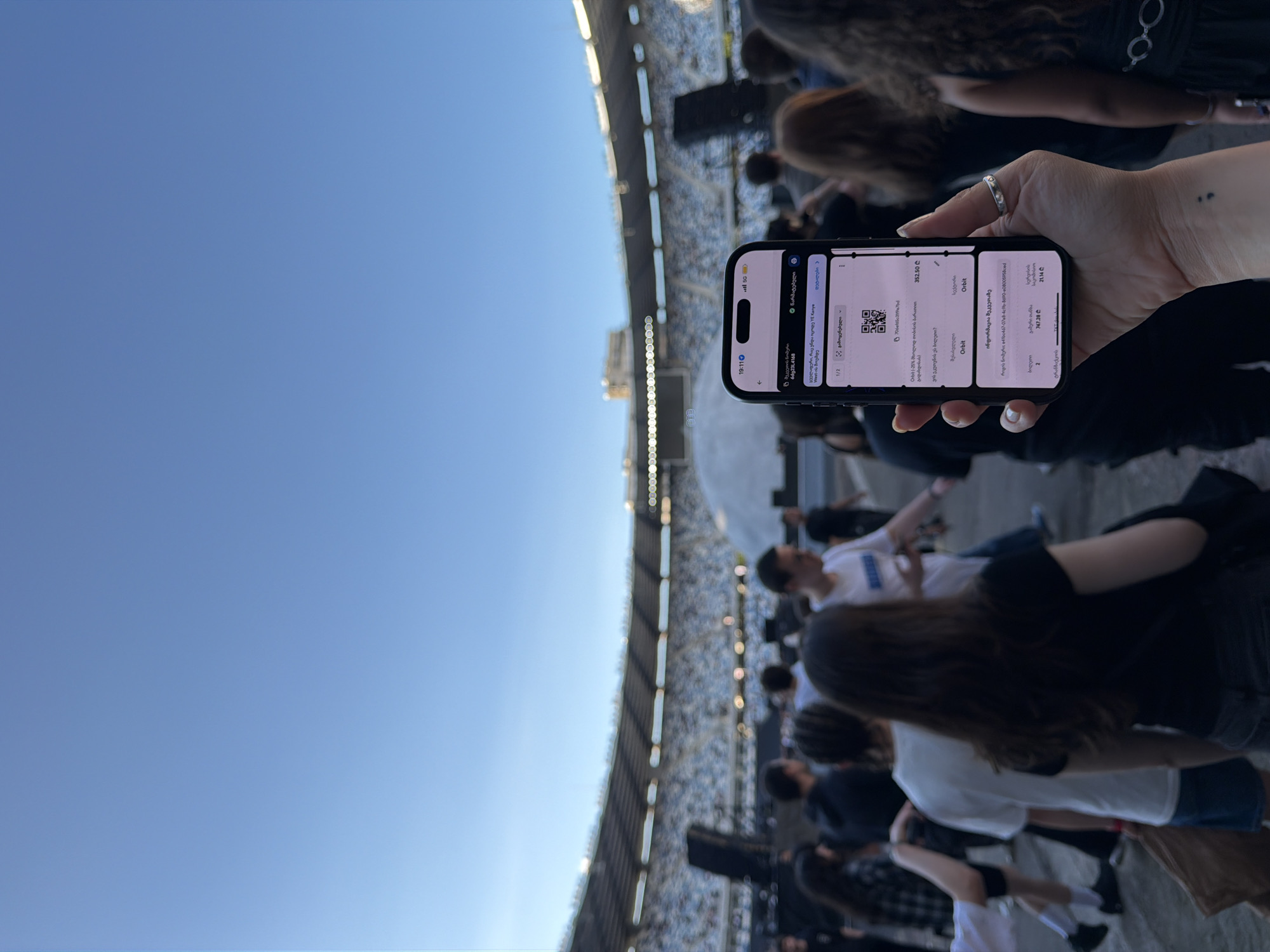 A person's hand holds a smartphone displaying a ticket for a YE Kanye West concert at the Dinamo Arena in Tbilisi, Georgia. The phone screen shows the ticket details, including a barcode and event information. In the background, a large stadium is filled with spectators, and a crowd of people is visible walking towards the stadium. The sky is clear and blue.
