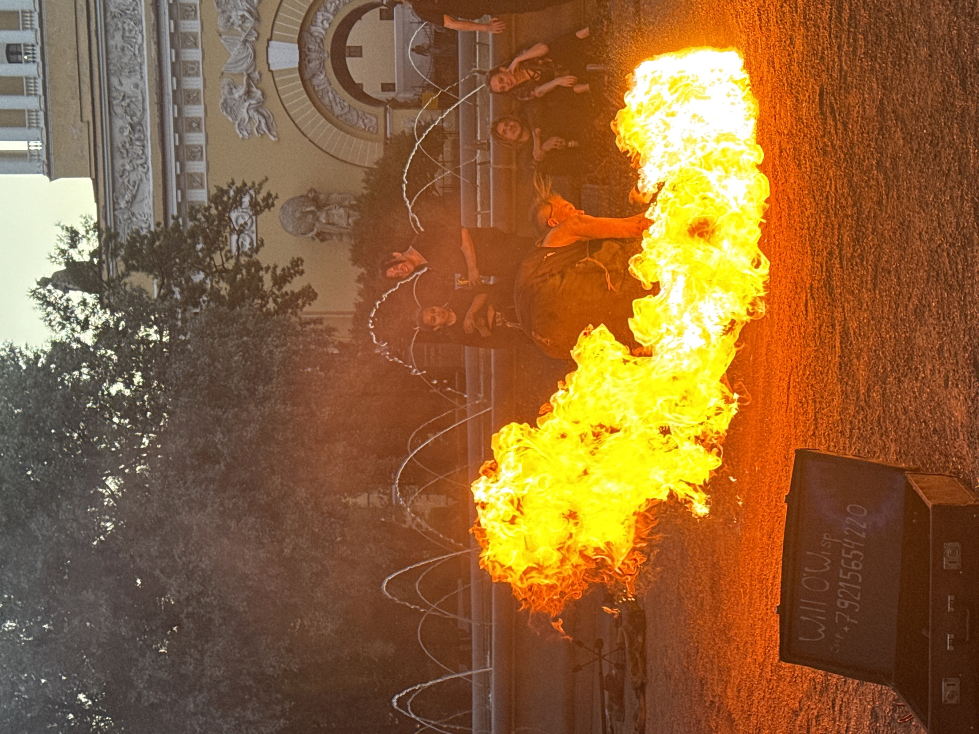 In front of the iconic yellow facade of the Admiralty Building in Saint Petersburg, Russia, a group of people are gathered. The foreground is dominated by a large, intense fire. To the left, an open, dark-colored suitcase is placed on the ground, with white lettering on its surface that reads "WiII O'Wisp" and "+79215654220". Several individuals are visible in the background, with one person in the center appearing to be manipulating the flames or a prop. Behind them, a series of arched fountains are spraying water. The architecture of the Admiralty Building, with its classical columns and decorative friezes, is visible in the upper background. The overall scene suggests an outdoor performance or street art display.
