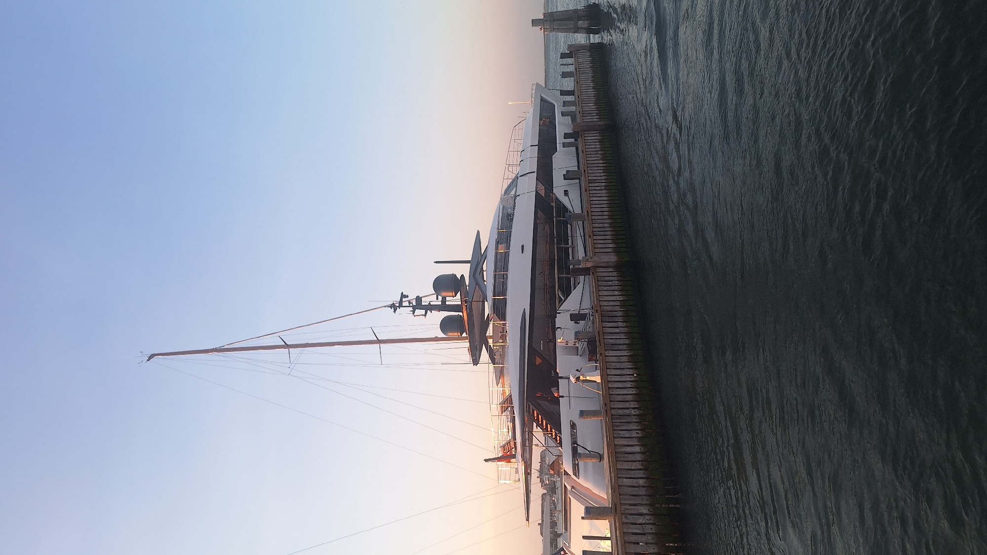 A large, white motor yacht with a prominent mast is docked at a wooden pier in the waters of Nantucket, Massachusetts. The yacht is sleek and modern, featuring multiple levels and dark accents. The sky above is clear and pale blue, transitioning to a soft pink near the horizon, indicating either sunrise or sunset. The water is dark and rippled. Several wooden pilings are visible along the pier, and a single, weathered piling stands in the water to the right of the yacht. The overall scene is serene, depicting the yacht moored in a harbor environment.