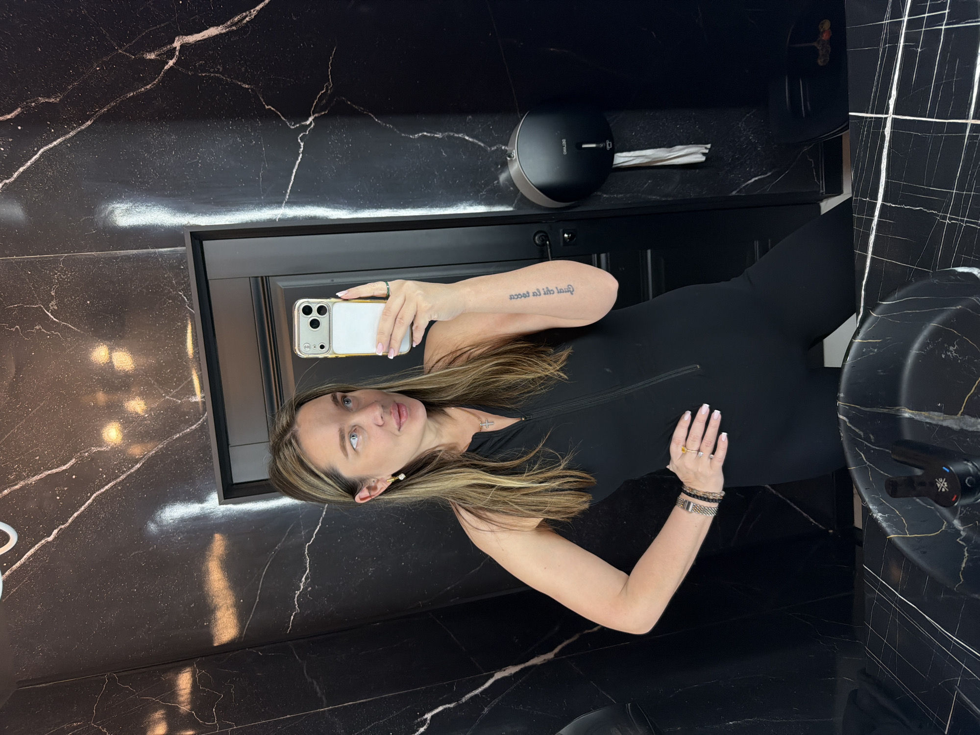 A woman is taking a selfie in a bathroom with black marble walls and fixtures. She is wearing a black jumpsuit. The bathroom features a sink, a mirror, and a toilet paper dispenser. No information is available to confirm that the location is in Moscow, Russia.