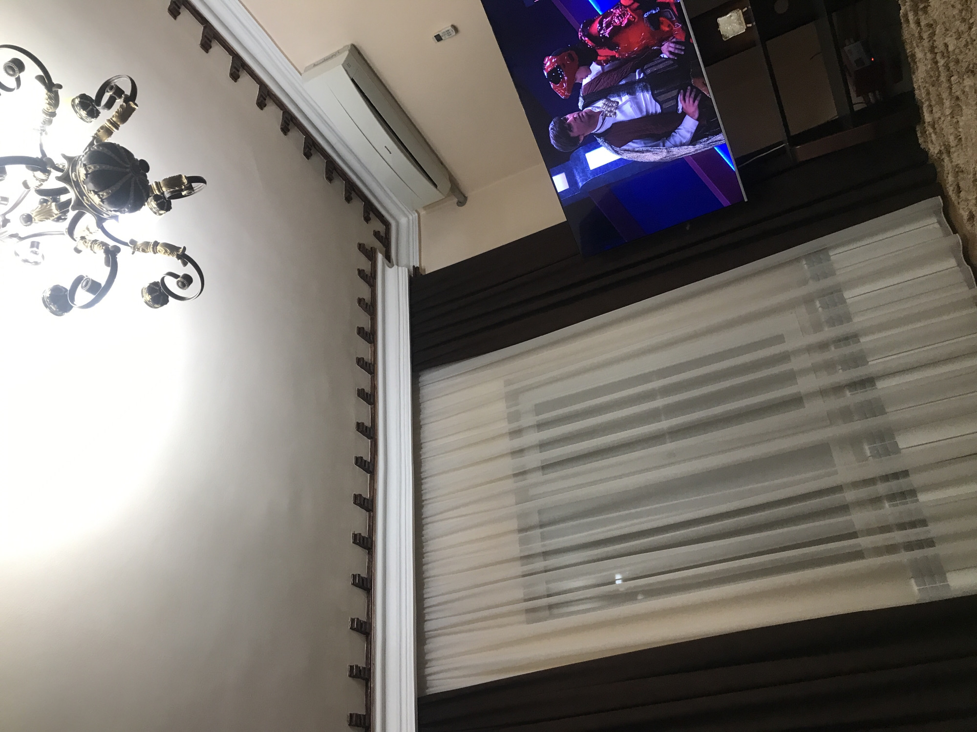 The scene appears to be an interior shot of a room in Yerevan, Armenia, featuring architectural details and furnishings. Dominating the left side is a white wall decorated with a brown, patterned molding. A decorative chandelier hangs from the ceiling. A split AC unit is mounted near the top of the wall, adjacent to a flat-screen television displaying a video or image. The right side of the scene includes a large window with sheer blinds, partially obscured by brown curtains at the top and bottom. The overall impression is that of a comfortable living space within a residential setting.