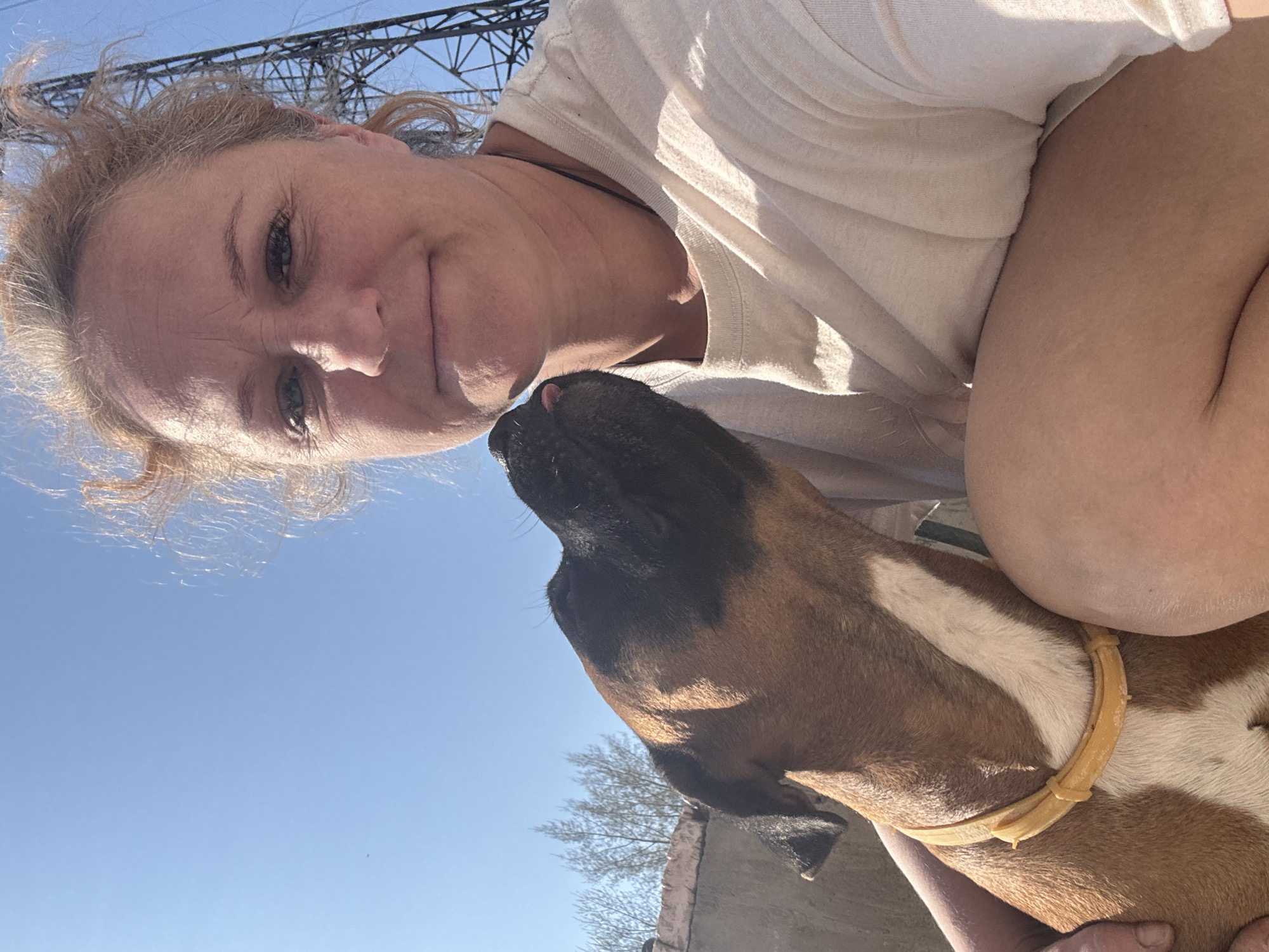 A woman with fair skin and blonde, curly hair poses for a selfie with a brown and white Boxer dog. The woman is wearing a light-colored t-shirt and has a slight smile. The dog is looking up at the woman and appears to be licking her face.
In the background, the sky is clear and blue. Power lines and a metal utility tower are visible in the upper left corner of the frame. A brick wall is partially visible along the bottom edge, indicating an urban or residential setting. Based on the location provided, this scene takes place in Chaikovsky, Russia.