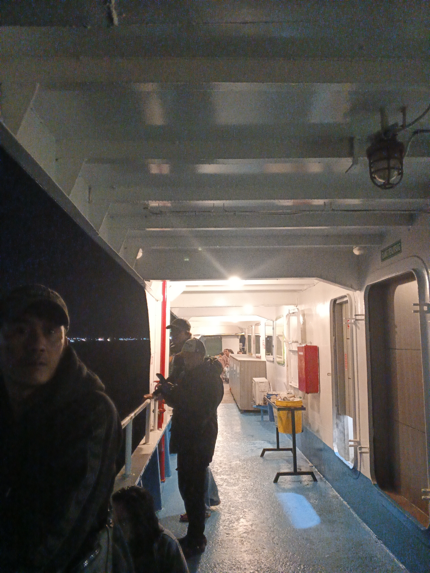 The image shows the interior of a ferry in Banyuwangi, Indonesia, likely at night. In the foreground, a person wearing a dark hooded jacket and a cap is looking towards the camera, their face partially obscured. To their right, another person in a dark jacket is standing, facing away. Further down the corridor, several other individuals are visible, some appearing to be passengers. The ferry's interior features white walls, a blue-painted floor, and industrial-style lighting. A red fire extinguisher is mounted on the wall. Outside the ferry, a dark expanse suggests it is sailing on water, with distant lights indicating a shoreline. The overall scene depicts a typical ferry environment with passengers in transit.