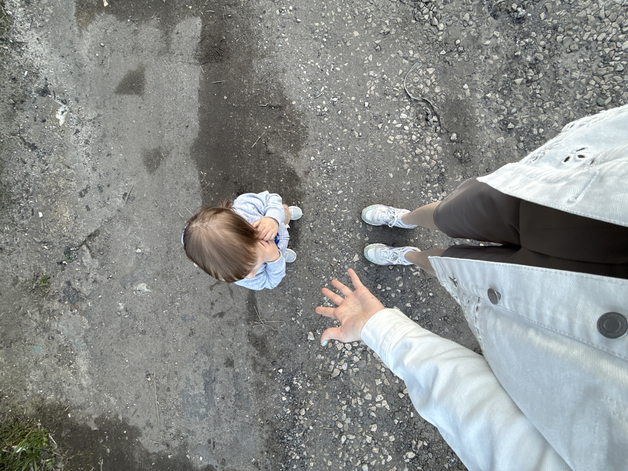 An overhead shot depicts a child, around 2 years old, dressed in a light blue top and darker blue bottoms, looking up. A person, presumably an adult, is standing nearby, wearing grey shoes, dark brown shorts, and a white denim jacket. Their hand is outstretched. They are standing on a coarse gravel surface. The location cannot be determined with the given information.