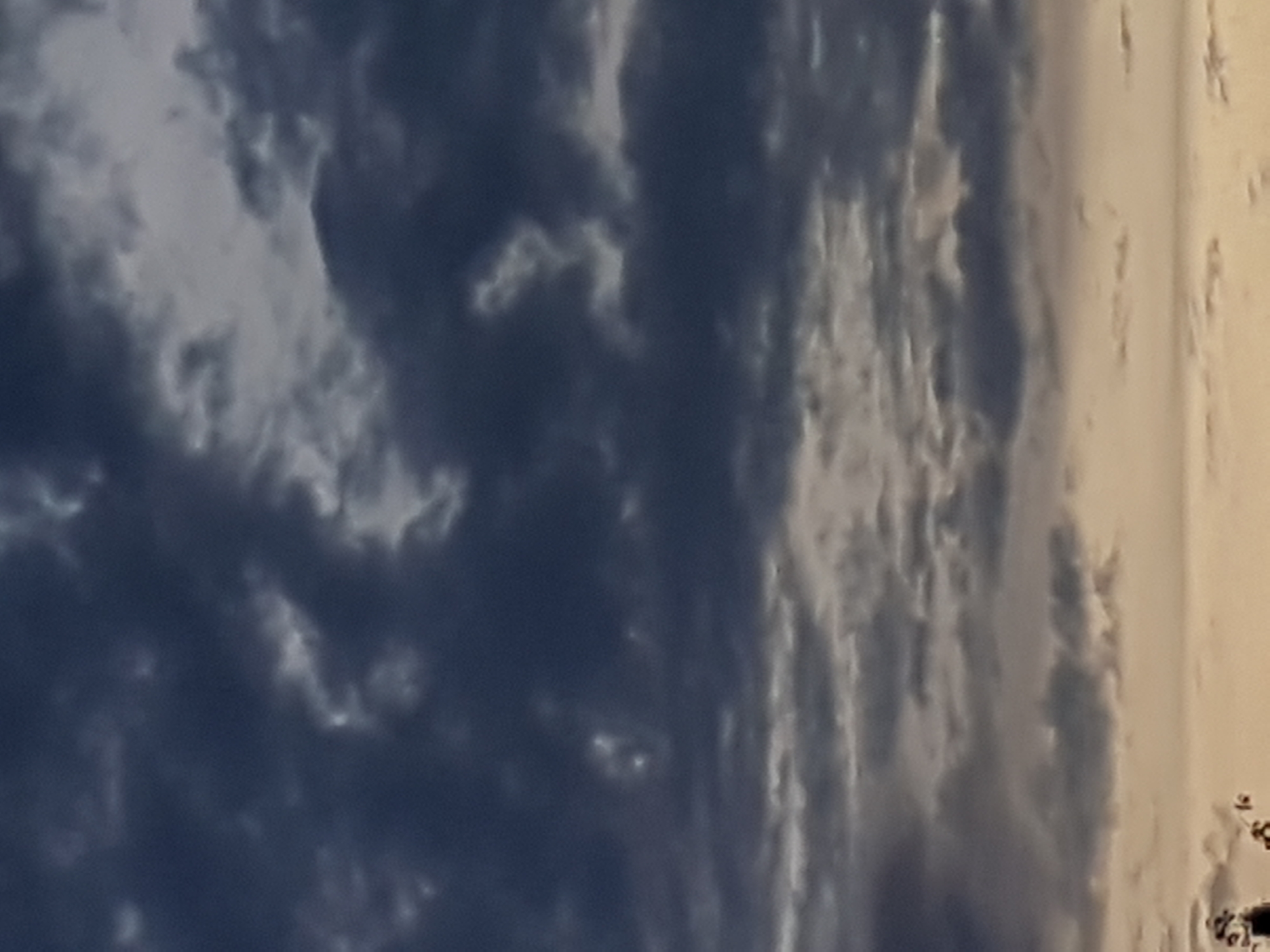 The image is a low-angle, medium shot of a cloudy sky at dusk. The clouds are dark and heavy, with some lighter patches of gray and white. The bottom of the image shows a faint hint of a golden-orange horizon, suggesting the sun has recently set. There are no people, subjects, or objects clearly identifiable in the sky. The lower left corner shows a small portion of dark foliage from what appears to be trees. The location is unspecified.