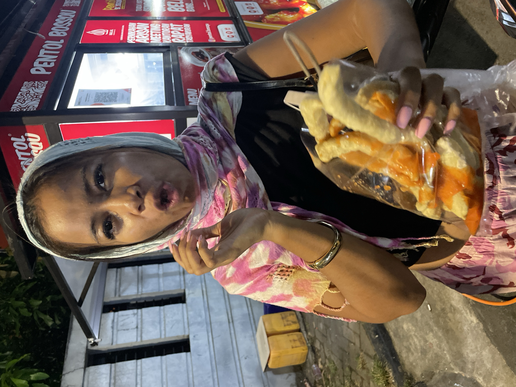 The image is a low-angle shot of a woman holding a plastic bag containing what appears to be food, possibly fries coated in sauce. She is positioned in front of a food stall labeled "PENTOL BOSSON," with additional text in Indonesian related to events such as weddings, meetings, and gatherings.
The woman is wearing a headscarf with a pink and white patterned design, a black top, and a pink skirt or pants. She has a gold bracelet on her wrist and light pink fingernails. She is looking at the camera with a playful expression.
The food stall has menus and signs visible. The overall scene appears to be outdoors, possibly at night, in an urban setting in Central Jakarta, Indonesia, given the text on the food stall. There is a concrete floor visible in the lower right.