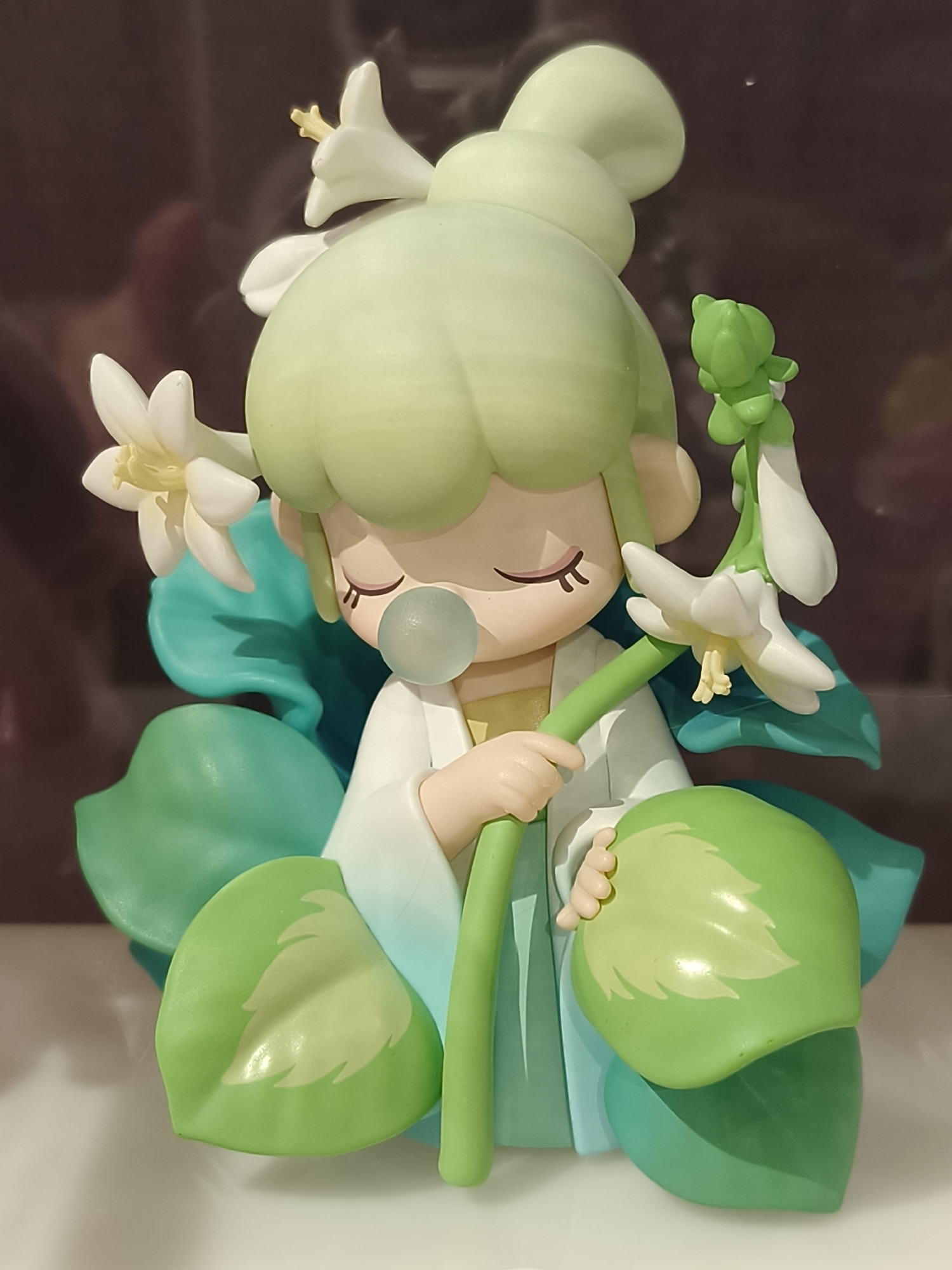 The image displays a stylized figurine of a youthful, human-like character, possibly a nature sprite or fairy. The figure has prominent, light pastel green hair styled in a large bun and bangs. Its eyes are closed with delicate dark eyelashes, conveying a serene or peaceful expression, and a translucent bubble is depicted near its mouth, resembling blown bubblegum.
The figurine wears an outfit that is predominantly white, with a subtle gradient transitioning to a light blue-green color towards the lower part. It is surrounded by and seated amongst numerous large, vibrant green leaves of various shades, from a light lime green to a deeper teal. Some of these leaves feature lighter green, feathered patterns.
Several delicate white flowers, each with a pale yellow center, are incorporated into the design. Two flowers are nestled in the figure's hair, and another cluster of flowers on a green stem is gently held in its hands. A small, bright green, bud-like creature or element is perched on one of the flower stems. The figurine's smooth, matte texture suggests it is crafted from plastic or resin. No visible text is present in the image.
The overall presentation is whimsical and tranquil, evoking a connection to nature. This figurine is located in Dnipro, Ukraine.