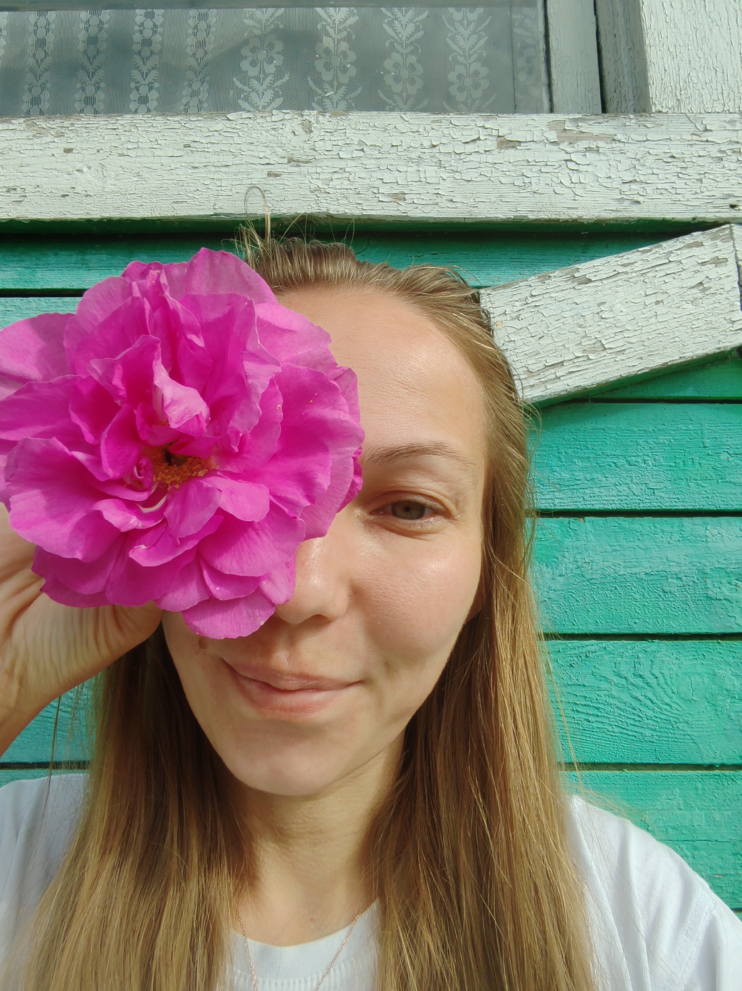 A young woman, with long blonde hair, is shown in a close-up, self-portrait, holding a large pink flower over her right eye. She is wearing a white t-shirt and a delicate necklace. The background is a weathered, teal-colored wooden wall with a white trim and a window with a lacy curtain. The image appears to have been taken outdoors during the daytime. Based on the architectural style of the window and the rustic wooden siding, this scene is likely located in a rural or suburban area of Чухлэм, Russia.