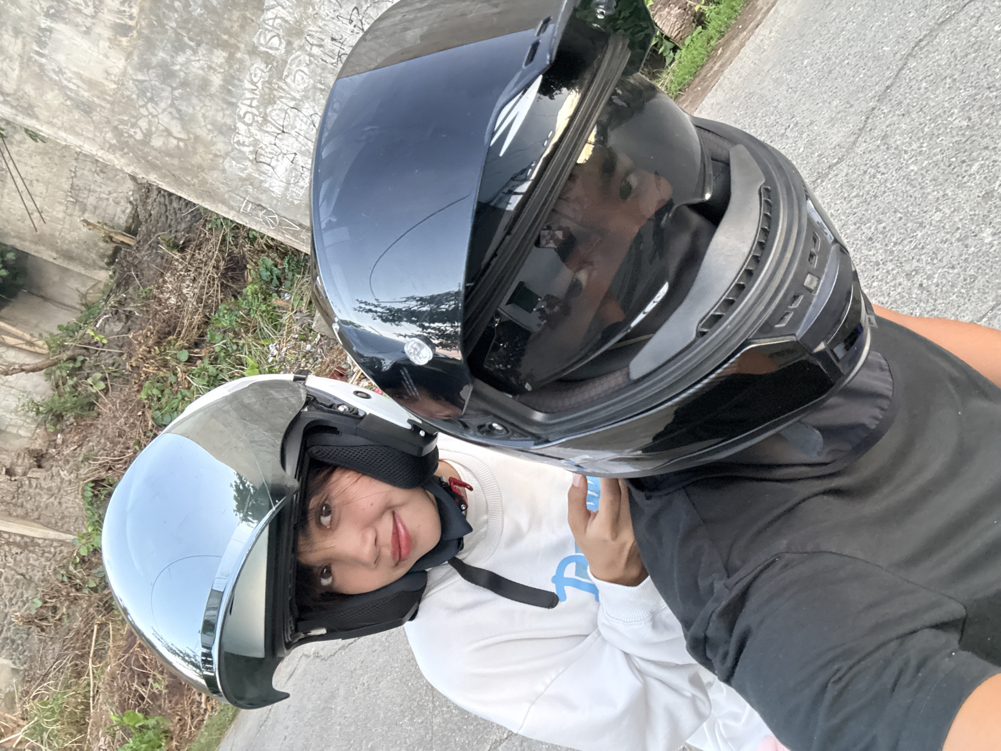 This is a selfie taken from a motorcycle, likely in Davao City, Philippines. Two individuals are wearing helmets. The person in the foreground is wearing a black full-face helmet, with their head and shoulders visible. The visor reflects a person's face and the surrounding environment, showing trees and possibly buildings. The person behind them is wearing a silver open-face helmet. This person has dark hair and is looking at the camera with a slight smile. They are wearing a white long-sleeved shirt with a blue logo. The background shows a dirt embankment with vegetation and a concrete structure with graffiti. The road surface is asphalt. The overall lighting suggests it is daytime, possibly late afternoon due to the warm tones.