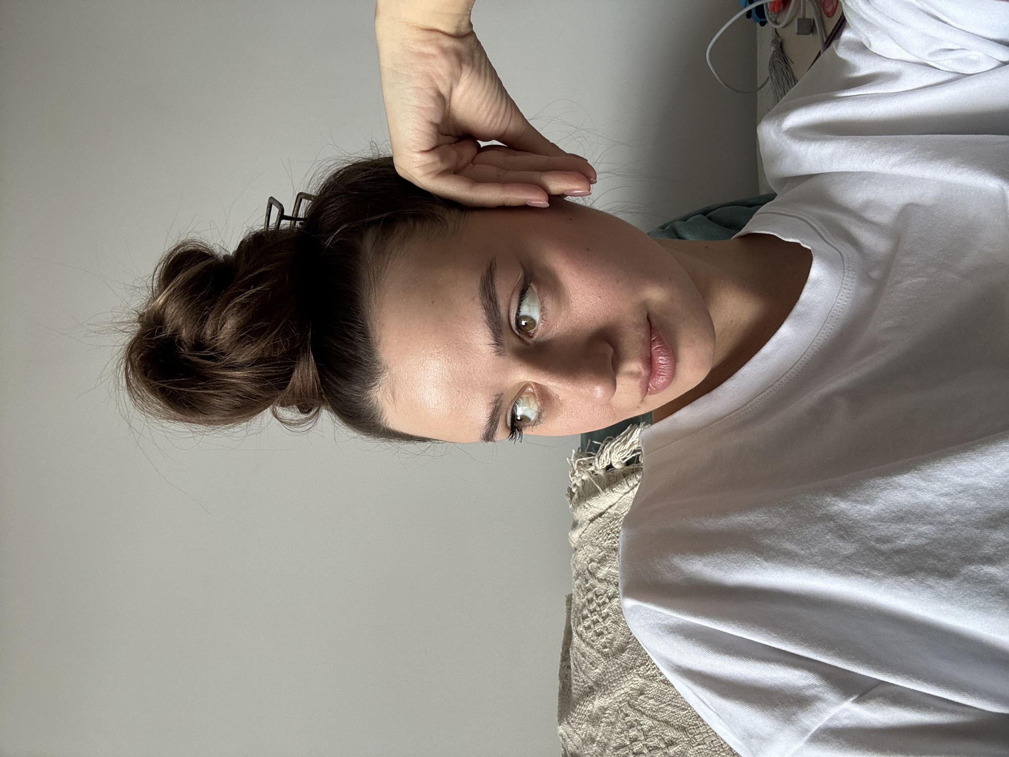 A woman is positioned indoors, angled toward the viewer. Her brown hair is styled in a bun, secured with a hair clip. She is wearing a white t-shirt. One of her hands is raised near her face. A portion of a bed or furniture is visible behind her. Wires are visible in the background. The location is unknown.