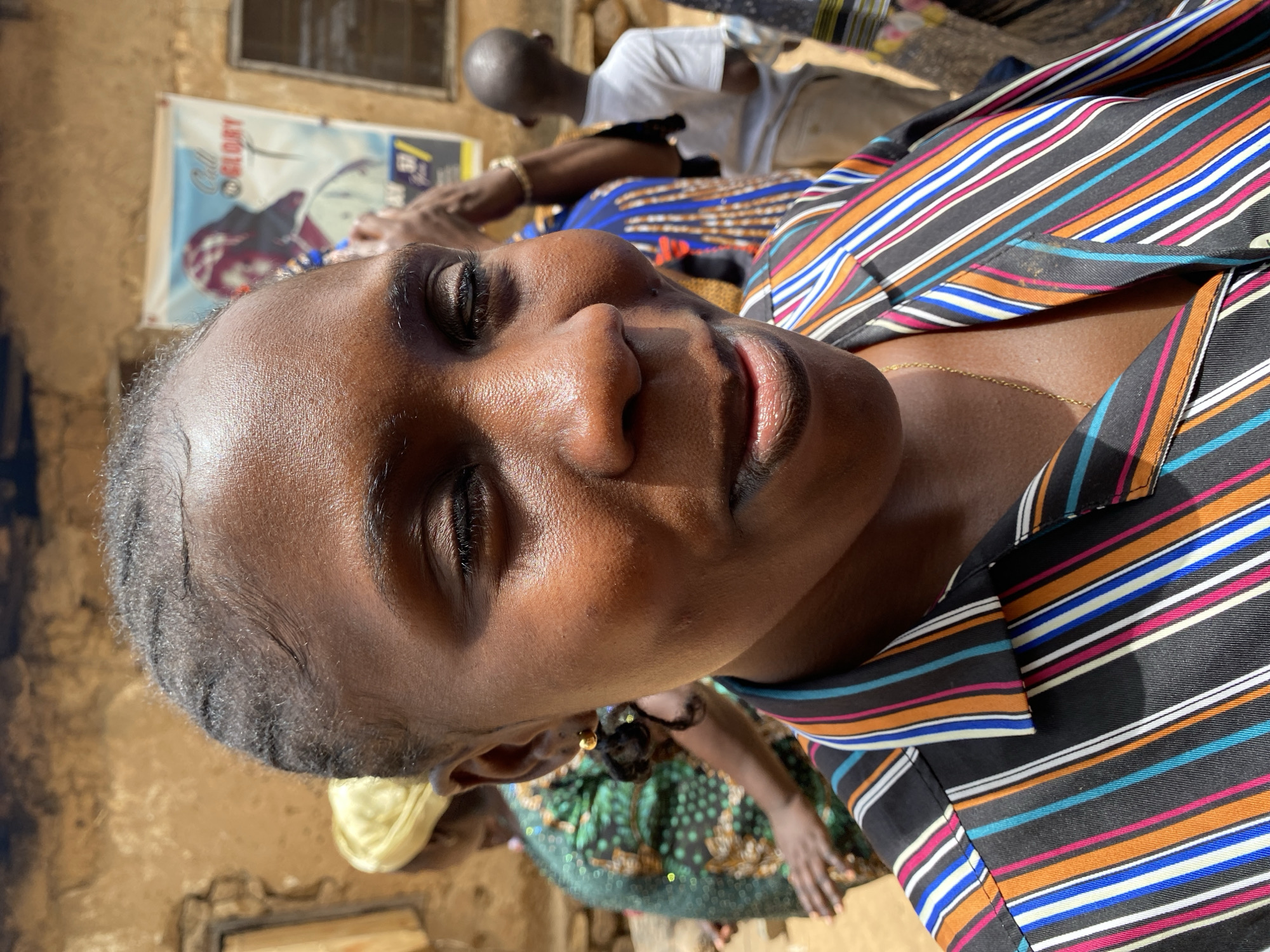 The image shows a close-up, low-angle shot of a dark-skinned woman with short graying hair wearing a striped shirt with black, orange, white, and blue lines. She has a gold earring and is wearing a gold necklace. Her eyes are closed, and her skin is illuminated by sunlight.
In the background, there is a partially visible bald man in a white shirt and beige pants. Behind him is another person wearing a blue shirt with yellow and blue patterns. Further back, a person is wearing a green patterned dress, and someone is wearing a yellow head covering.
A weathered wall with a poster hanging on it can be seen on the left. Based on the location requested, the scene is most likely outdoors in Vom, Nigeria.