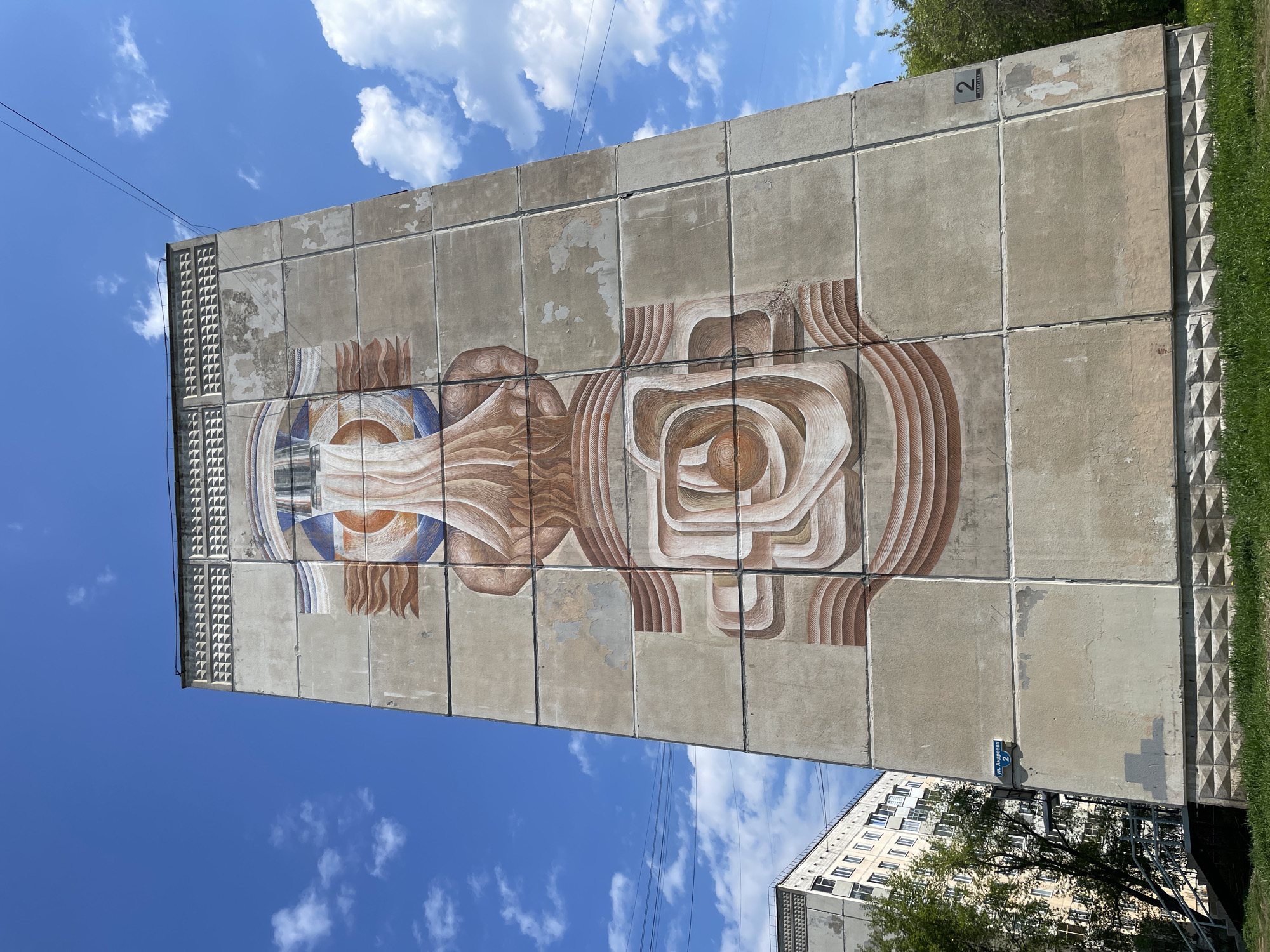A low-angle, outdoor shot shows a section of an apartment building with an artwork on its facade, situated in Zheleznogorsk, Russia. The building is constructed of square, concrete blocks with an abstract mural painted across the facade. The mural features patterns with curved lines in shades of tan, brown, orange, and blue, designed to create a visual depth.
On the right edge of the building, there is green vegetation, likely trees and grass. The number "2" is visible near the top right edge of the building. A blue street sign is also seen on the side of the building near the bottom.
The sky is bright blue with scattered white clouds. In the bottom right corner of the frame, there's another building with windows and geometric design elements, partially obscured by trees. Utility wires are visible.