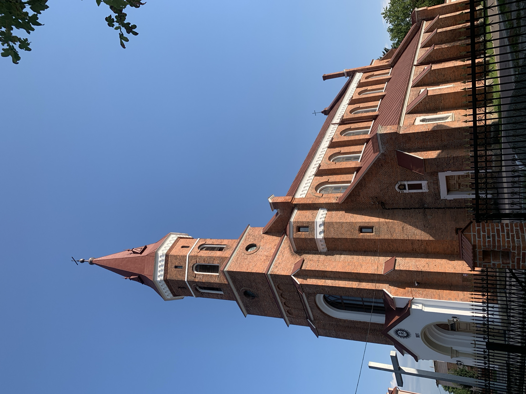 The image shows a brick church with a red roof, featuring a tall bell tower topped with a spire. A large white cross stands to the left of the church entrance, which has a white facade with the year "1897" visible. The church is enclosed by a black metal fence. The sky is clear and blue. The architectural style and the presence of the cross suggest it is a Christian place of worship. Based on available information, this is likely the Church of the Blessed Virgin Mary in Kolomyia, Ukraine.