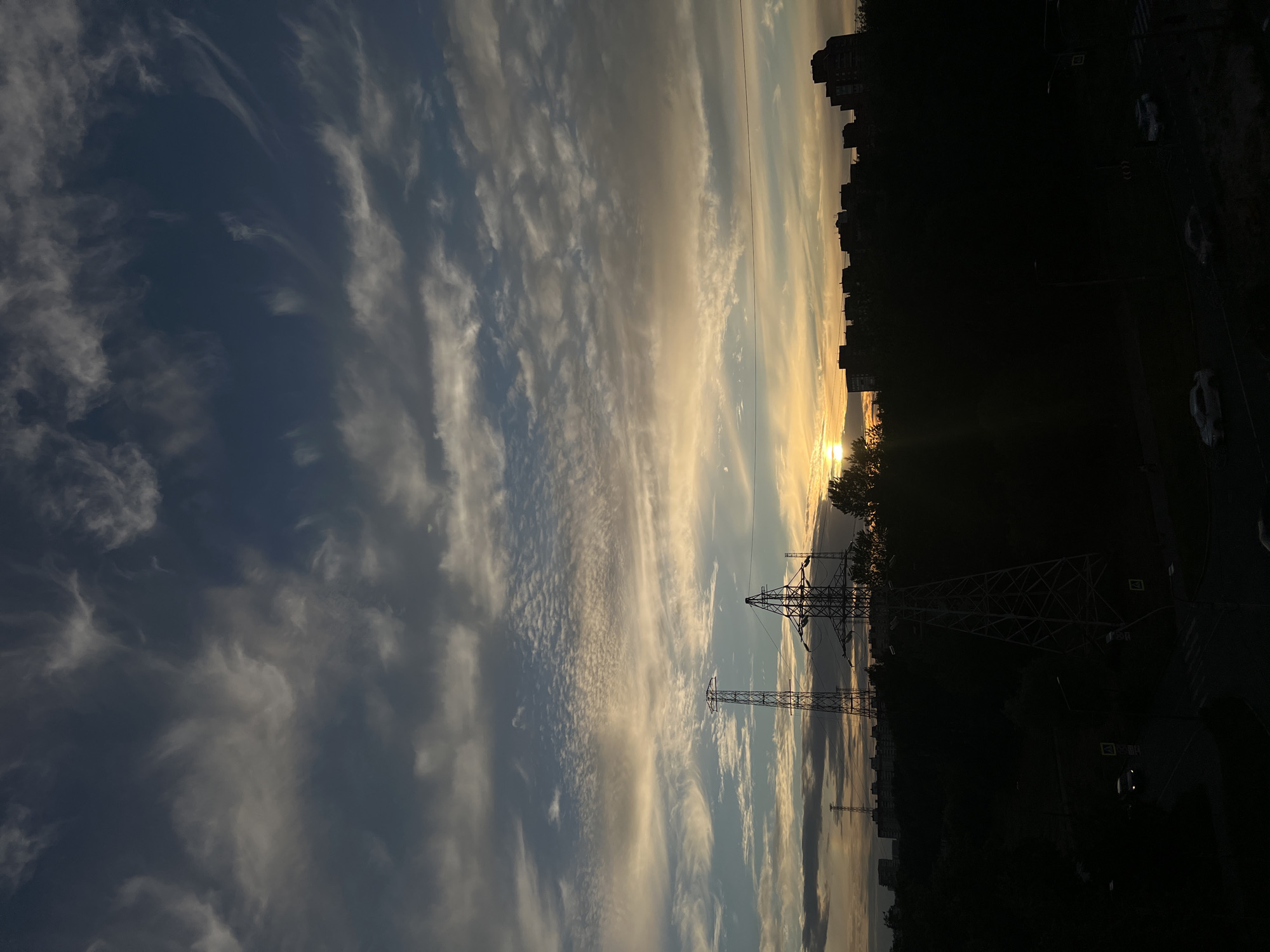 This is a vertical, low-angle, outdoor photograph taken during sunset. The sky is filled with clouds, ranging from dark grey to bright orange and yellow, indicating the sun is low on the horizon. The sun itself is visible as a bright disc near the center of the image. Silhouetted against the sky are several tall, lattice-like structures that appear to be electrical pylons. Beyond the pylons, a line of buildings forms a dark silhouette against the lighter sky, suggesting an urban environment. In the foreground, a dark, silhouetted landscape includes trees and a road with a few cars. The image captures a transitional time of day in what appears to be an industrial or residential area with significant infrastructure.