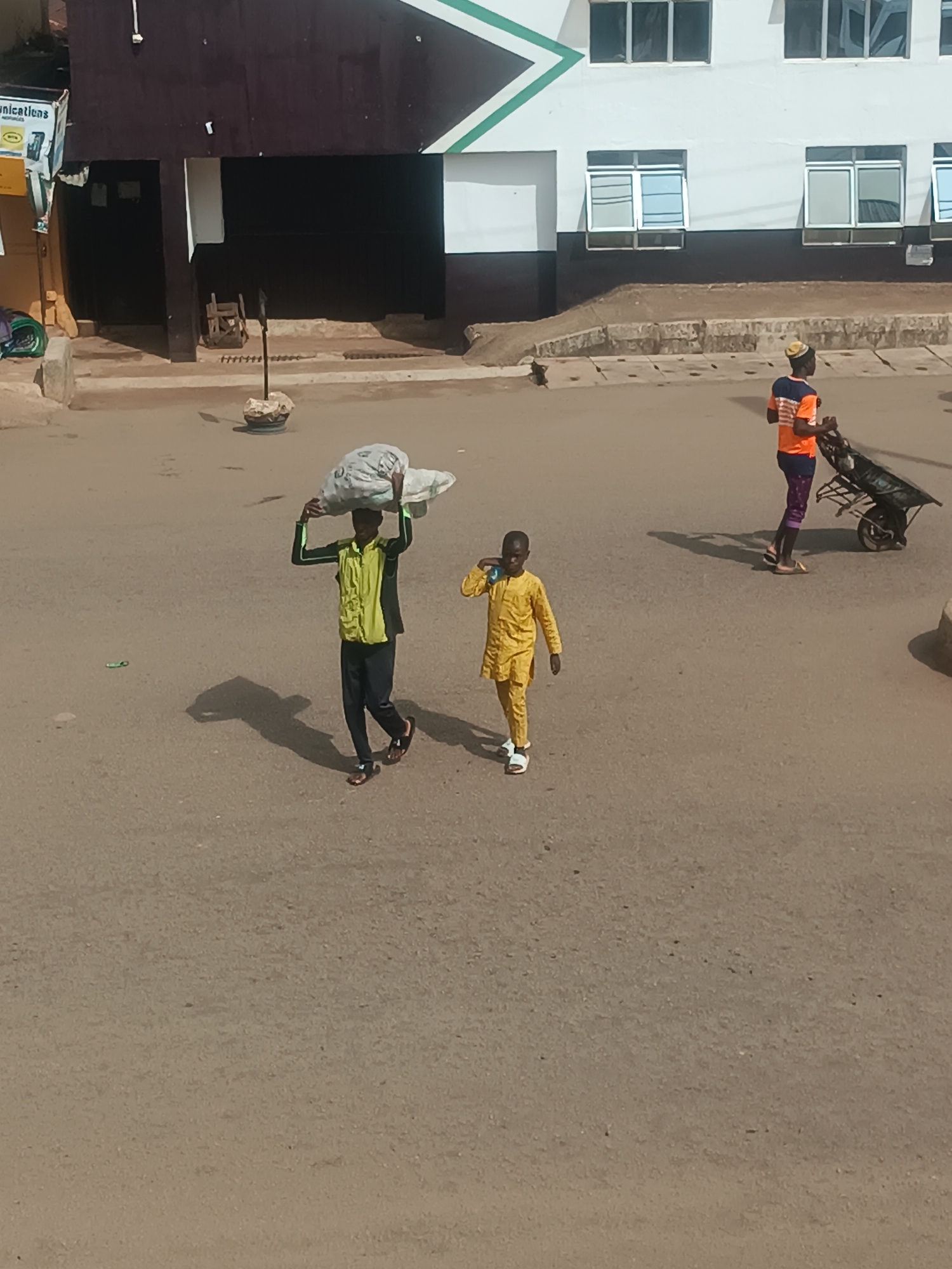 The image depicts a street scene in Bukuru, Nigeria. In the foreground, two young males are walking. One is carrying a large sack on his head and is wearing a green and black jacket over dark pants. The other, dressed in a yellow outfit, is walking beside him. In the background, a person wearing an orange shirt and patterned pants pushes a wheelbarrow. The backdrop includes a building with dark brown and white sections, multiple windows, and what appears to be a green chevron design. The street itself appears to be paved and there are a few other indistinct items visible in the area.