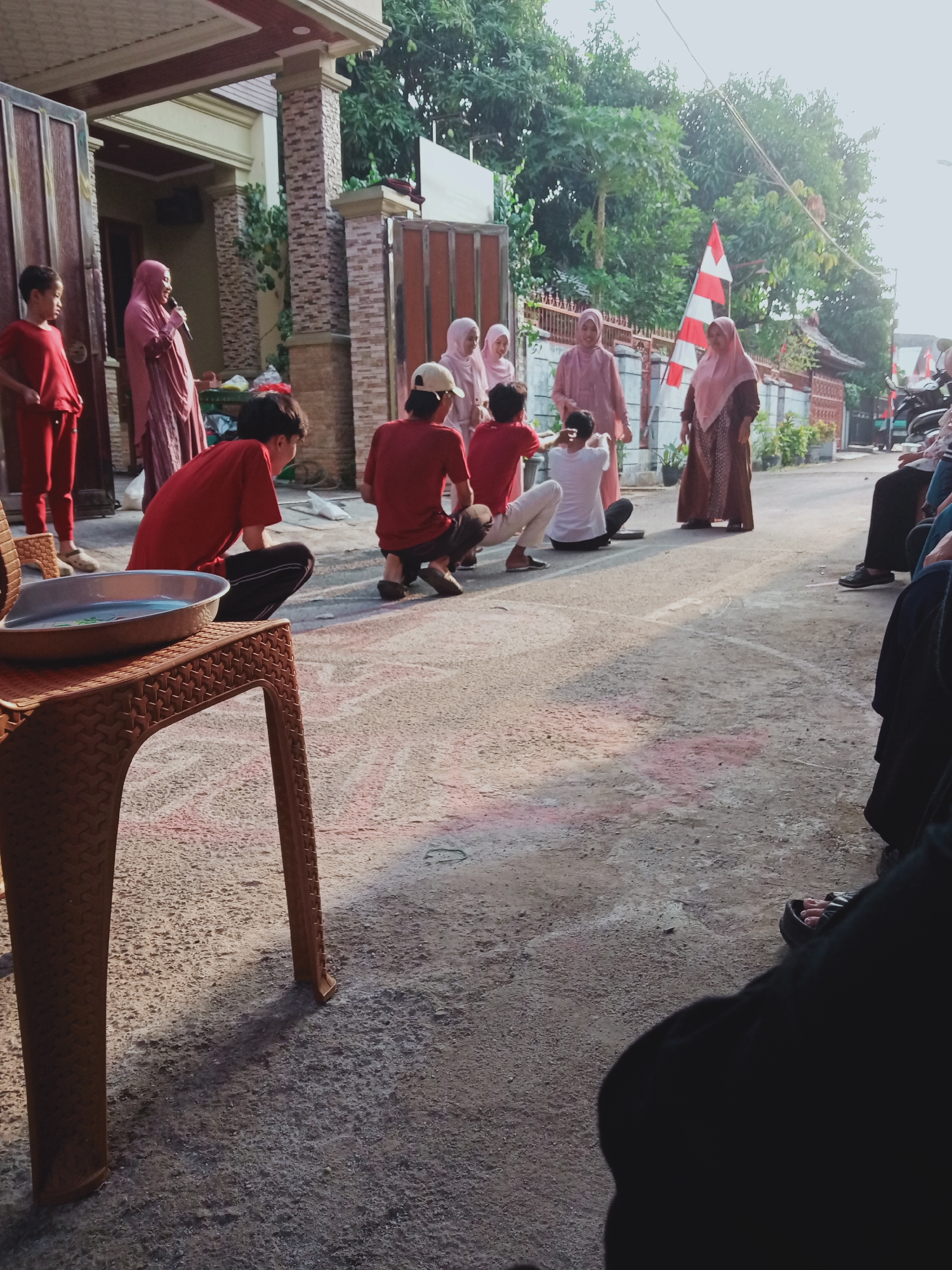 The image depicts an outdoor gathering of people, likely in a residential area, possibly on a street or in a communal space in East Jakarta, Indonesia. In the foreground, a brown plastic chair with a metallic bowl containing liquid sits prominently. The ground is paved with asphalt and shows chalk markings, suggesting a game or activity is taking place.
A group of individuals, predominantly wearing red and pink attire, are engaged in what appears to be a children's game or a community event. Several children in red shirts and pants are positioned on the ground, some crouching and others sitting. Adults, mostly women wearing hijabs and long dresses in shades of pink and brown, are observing or participating. One woman in a pink hijab is speaking into a microphone, potentially addressing the crowd or announcing something.
In the background, a red and white striped flag is visible, possibly indicating a national celebration or event. Buildings with beige and brown facades line the area, with trees and foliage providing a natural backdrop. The overall atmosphere seems to be festive and communal. The presence of motorcycles parked along the side suggests this is a commonly used thoroughfare or gathering spot. The lighting indicates it is daytime, likely late afternoon given the shadows.