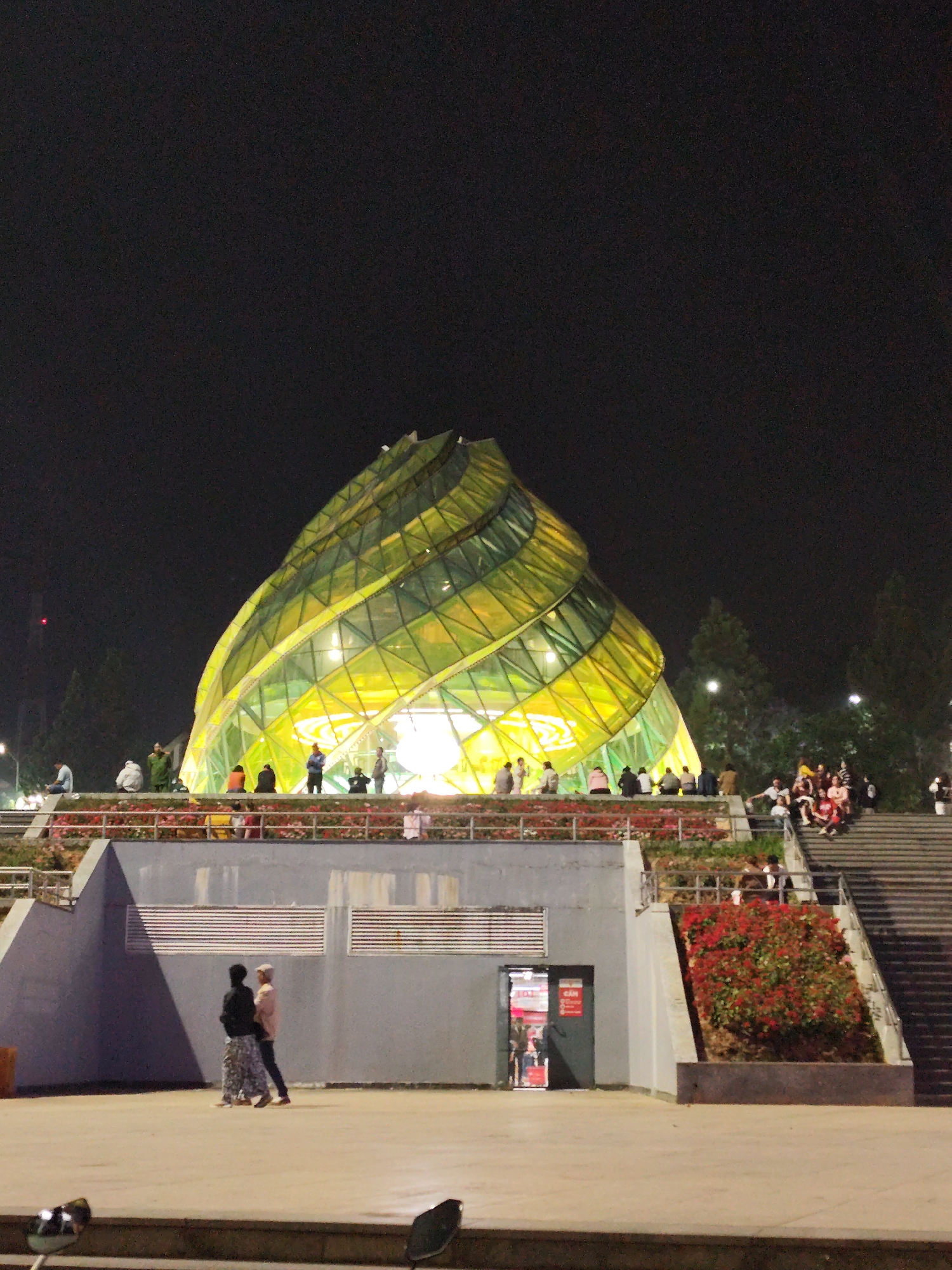 The image shows the Dalat Market in Da Lat, Vietnam, at night. The most prominent feature is the Artichoke Flower building, constructed from yellow and green glass, illuminated from within. People are present at the base of the structure, some sitting on a wall or railing. Below the building, a concrete structure with metal vents is visible, and two individuals are standing nearby. Stairs lead up to the building on the right side of the image, where more people are sitting. Bushes with red flowers grow along the base of the stairs. The overall scene is dark, with the Artichoke Flower building brightly lit and casting light onto the surrounding area. A door is visible on the right with a red poster. At the bottom left are a couple of unidentifiable dark objects.