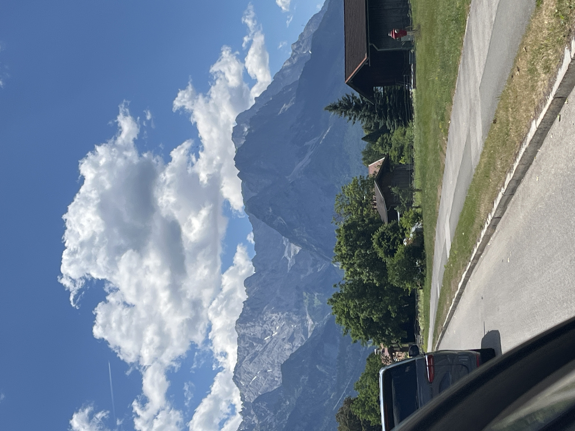 This image captures a scenic view of the Bavarian Alps, likely taken from a moving vehicle on a road in or near Garmisch-Partenkirchen, Germany. In the foreground, the edge of another vehicle is visible on the left, with its dark-tinted window obscuring much of its interior. Ahead, a dark grey minivan drives along an asphalt road with a paved pedestrian or bike path running parallel to it. The road is bordered by a grass verge.
In the mid-ground, a cluster of residential buildings with dark wooden facades and gabled roofs are nestled amongst lush green trees. A prominent red fire hydrant stands near one of the buildings. Evergreen trees and deciduous trees create a dense foliage backdrop.
Dominating the background are the majestic, rugged peaks of the Alps, their grey rocky surfaces showing patches of snow. The mountains are bathed in sunlight under a bright blue sky dotted with fluffy white clouds. A thin contrail from an aircraft is visible in the upper left corner of the sky. The overall impression is one of a clear, sunny day in a mountainous region with scattered human settlements.