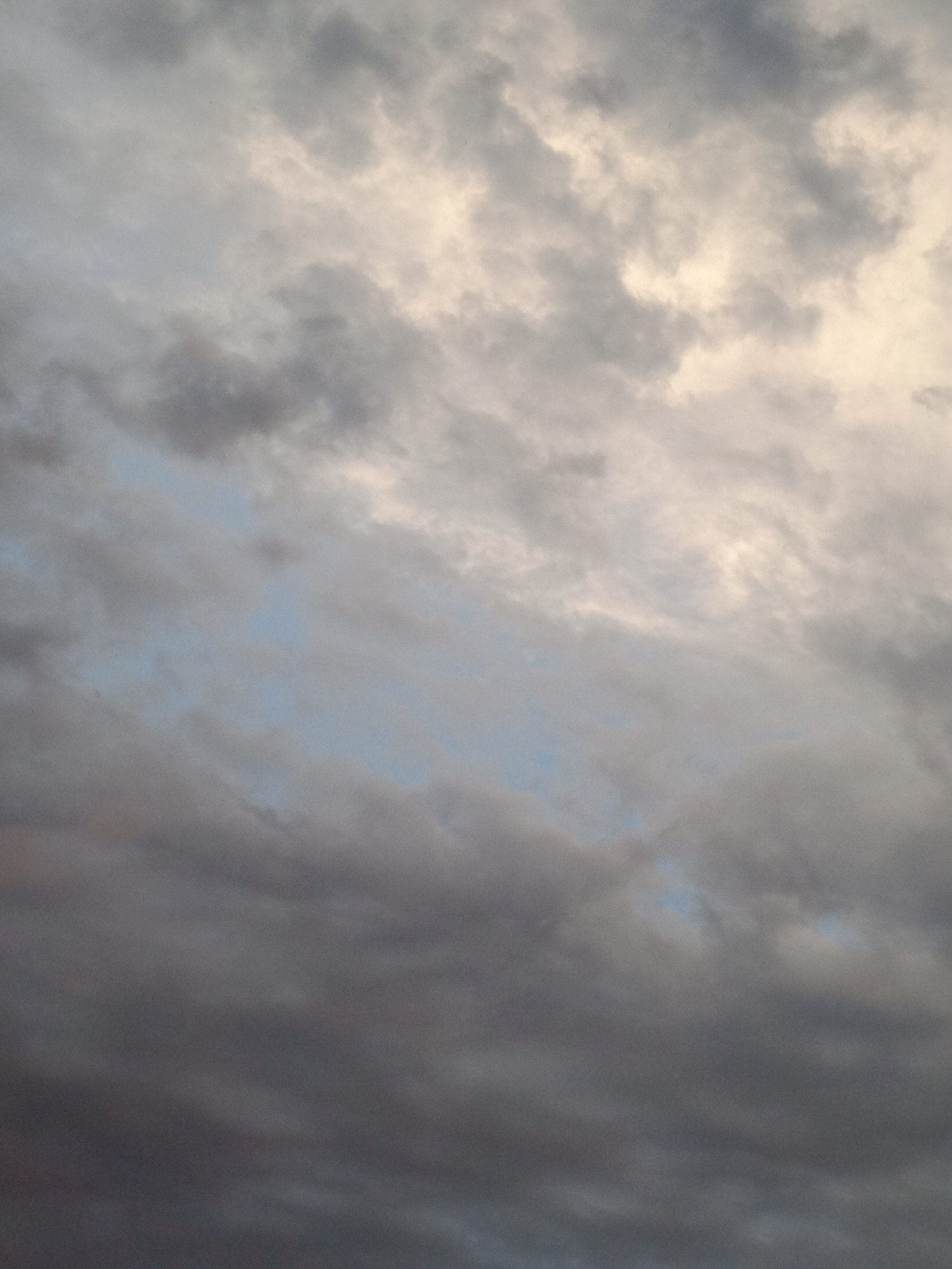 The image is a close-up, overhead view of a cloudy sky. The clouds are varied in color and texture, ranging from bright white and yellow near the top, to gray and dark gray lower down. There are hints of light blue sky visible between some of the clouds. The scene is devoid of any human presence or identifiable landmarks. The image appears to be a photograph of the sky taken from ground level, looking upwards. It is not possible to determine the specific location in Pankrushikha, Russia, from the image alone.