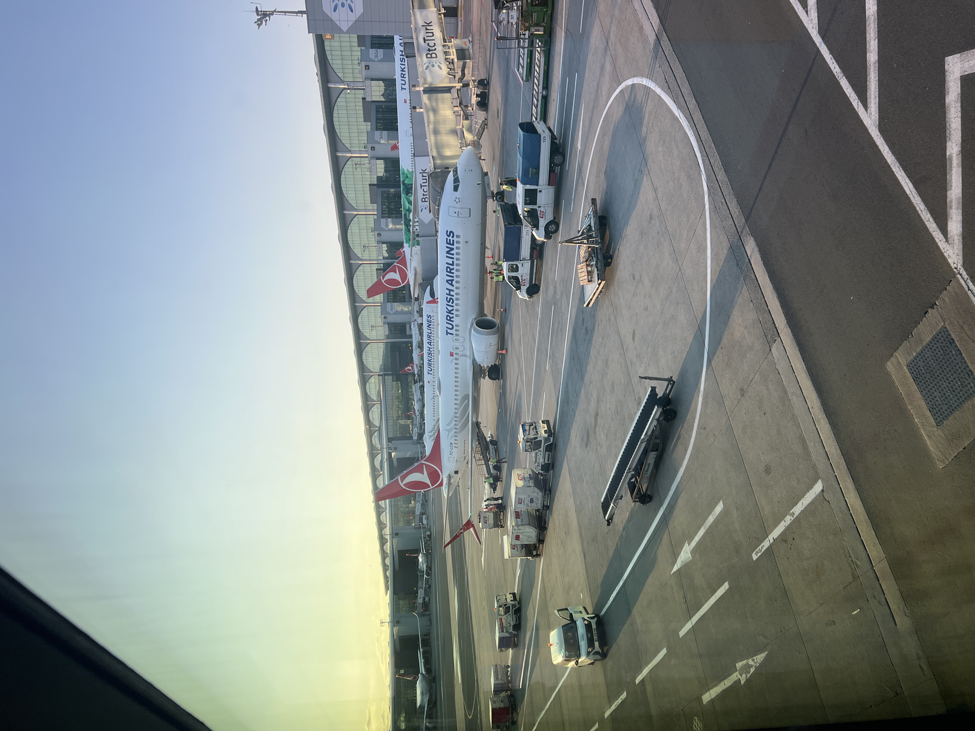 The image depicts an airport tarmac in Arnavutköy, Turkey, bathed in the warm light of what appears to be sunrise or sunset. Several Turkish Airlines airplanes are parked at gates, with their iconic red and white logos visible. Ground support vehicles, including baggage tugs, service trucks, and luggage carts, are actively engaged in operations around the aircraft. Workers in high-visibility vests can be seen attending to the planes and equipment. The terminal building, characterized by its large arched windows and modern architecture, forms the backdrop. The sky above is a gradient of pale blue and yellow, suggesting early or late day. The scene is a typical operational view of a busy airport.