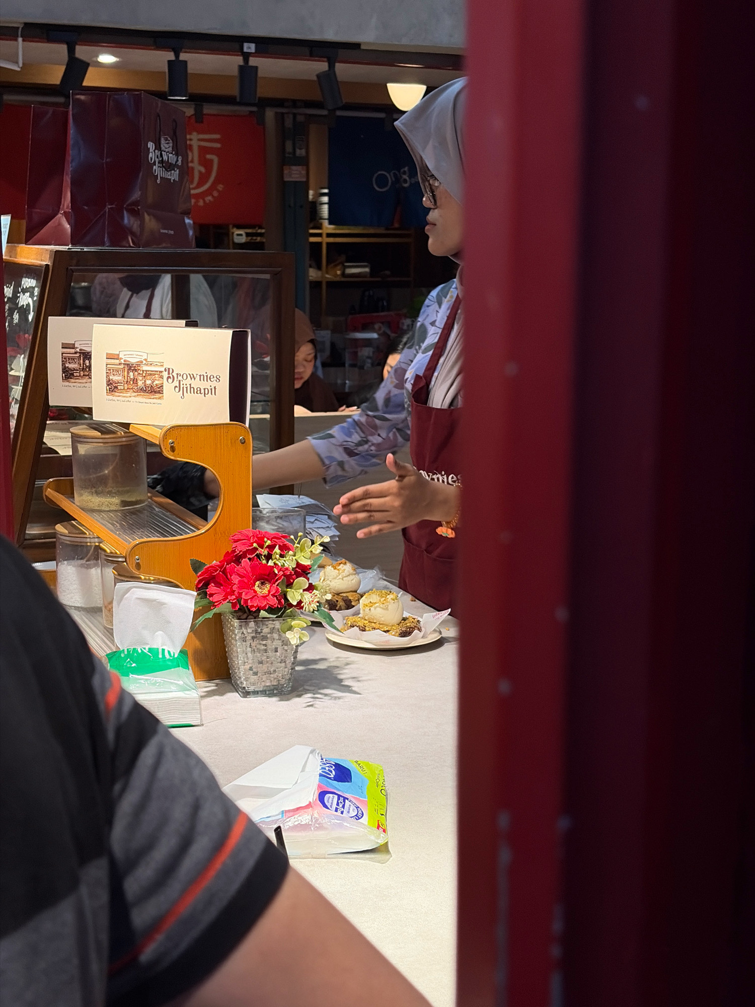 The image captures a scene at a food stall, likely in Bandung City, Indonesia, as indicated by the text "Brownies Djihapit" which is a popular Indonesian brownie brand often found in Bandung. A vendor wearing a gray hijab and a maroon apron is interacting with a customer whose arm and shoulder are visible in the foreground. The vendor is serving a dessert, possibly a brownie with toppings, on a white plate. In the background, there are various items typical of a food stall, including paper bags with the "Brownies Djihapit" logo, clear containers with ingredients, a flower arrangement in a decorative pot, and what appears to be a grater. The stall is brightly lit, with shelves and a display case visible. The overall atmosphere suggests a commercial setting focused on food service.