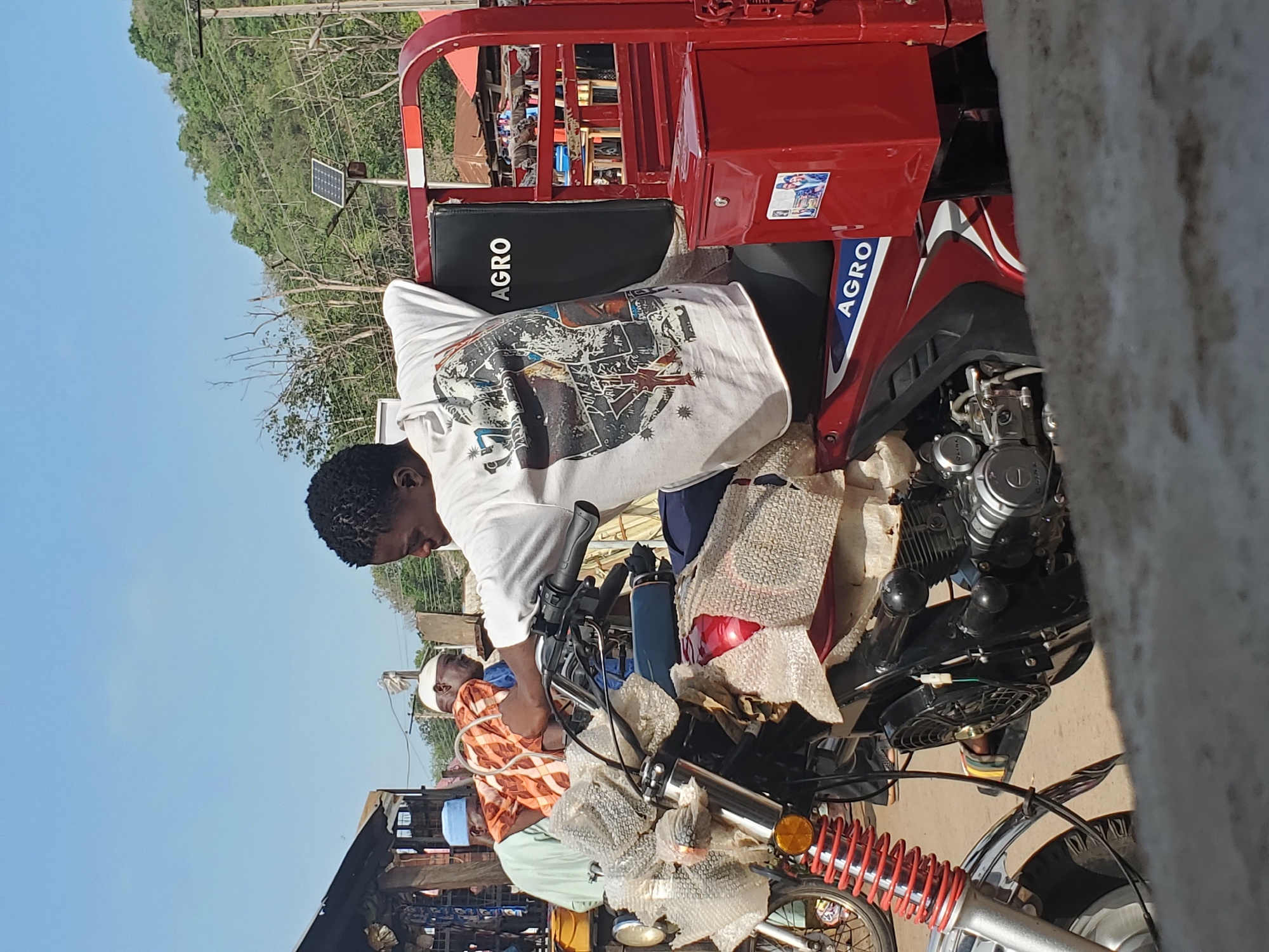 This image captures a vibrant daytime scene in Mararaba, Nigeria, featuring people and motorcycles under a clear blue sky.
The central focus is a red three-wheeled motorcycle, commonly known as a tricycle or keke napep, which dominates the mid-ground. A young man, wearing a white t-shirt adorned with a complex graphic design, is leaning over the front handlebars and engine area of this red tricycle, appearing to be working on it or inspecting it. The tricycle prominently displays the word "AGRO" in white letters on its black seatback and again in white and blue on its red side panel.
Next to the red tricycle, another motorcycle is visible, featuring red suspension springs. Several components of this motorcycle, including parts of its exhaust and other accessories, are wrapped in protective bubble wrap, suggesting they are new, recently delivered, or awaiting assembly. A blue water bottle is also seen on one of the bikes.
In the background, another individual, possibly older, is partially visible, wearing a patterned orange and white garment and a white cap, standing near other vehicles. The environment behind them reveals a green, tree-covered hillside and a solar panel mounted on a pole, indicating an outdoor, possibly semi-urban or roadside setting. The bright lighting suggests it is mid-day.
Visible text in the image is "AGRO," appearing on the seat and side of the red tricycle.
