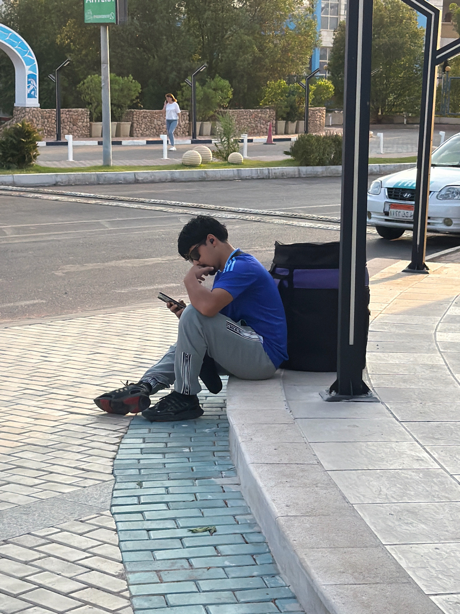 A young man in a blue shirt and grey pants sits on a raised concrete ledge, looking down at his phone. He wears sunglasses and dark sneakers. To his right is a large black and purple bag. In the background, a white car is parked, and a woman in a white shirt and jeans walks past in the distance. The scene appears to be on a street or promenade in Sharm El Sheikh, Egypt, with trees, lampposts, and decorative stone walls visible. The ground is paved with light-colored tiles and some blue-green tiles.