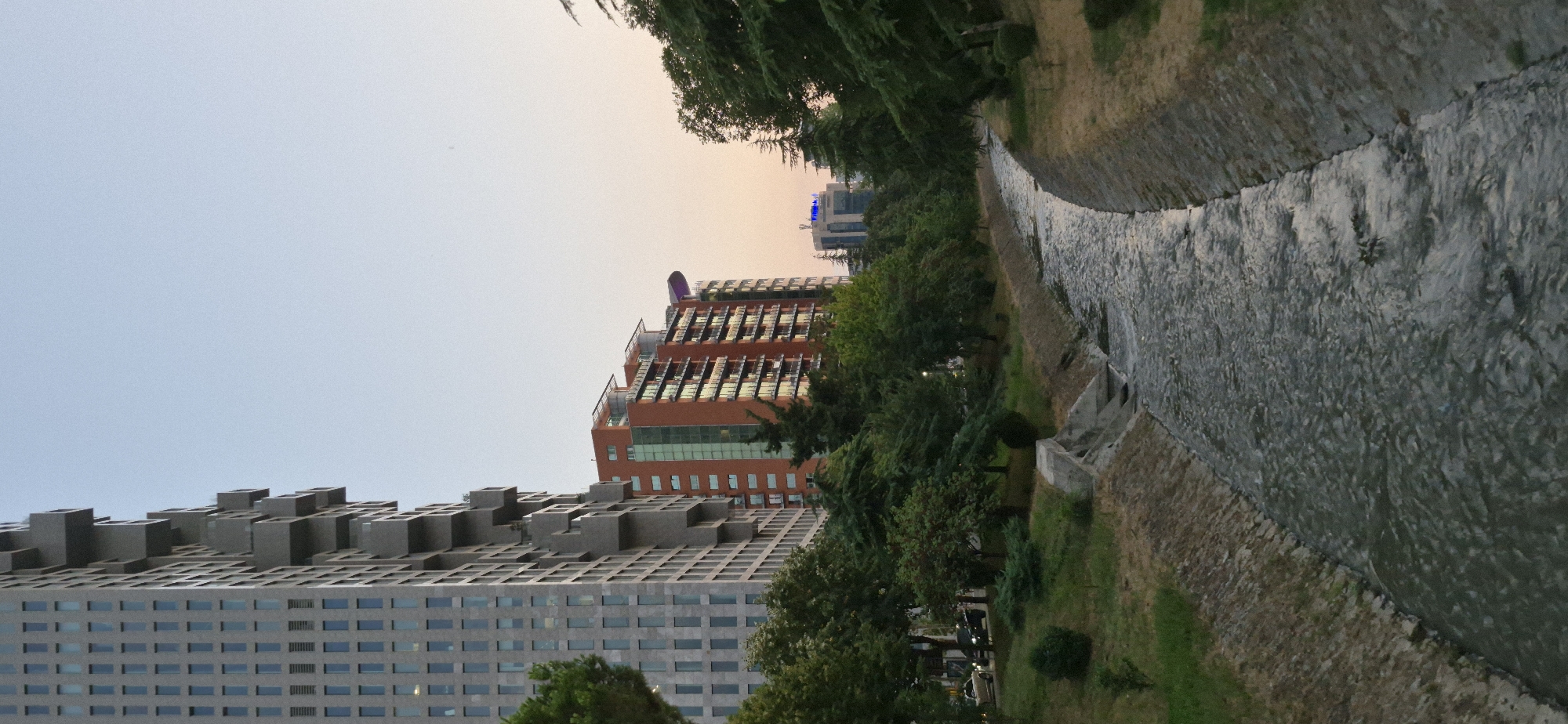 The image shows a man-made canal running through a park-like area in Tirana, Albania. On the left, a tall, modern building with a grey facade and protruding balconies dominates the skyline. To its right, a shorter, reddish-orange building with multiple balconies and large windows reflects the ambient light. Further in the background, another building with blue illuminated signage is visible. The canal, lined with stone and concrete embankments, has a stream of water flowing through it. The banks are covered with grass and trees, with some bushes and smaller trees planted near the canal. The overall scene suggests an urban environment with green spaces integrated into the cityscape. The lighting indicates it is likely late afternoon or early evening. There are no people visible in the image.