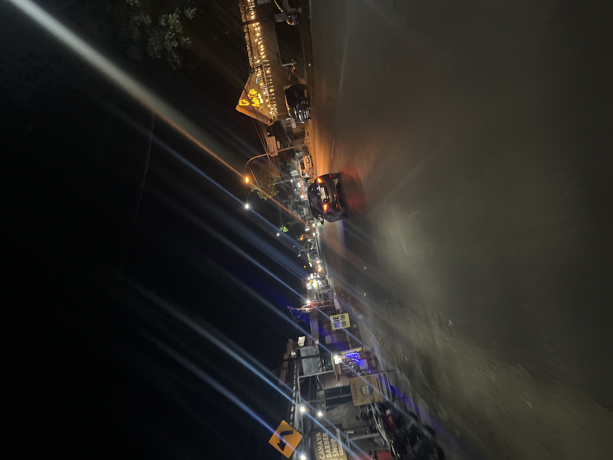 The image depicts a nighttime scene on a street in Kalinyamatan, Indonesia. A dark-colored sedan is driving down the road, with its taillights illuminated. To the left of the road, there are several businesses, including a barbershop with signage that reads "CAHAYA BARBER SHOP." There are also motorcycles parked along the sidewalk and a yellow directional sign indicating a left turn. Further down the road, on the right side, is a building with a pitched roof, adorned with numerous lights, appearing to be a cafe or restaurant, with the name "Anitre Cafe" visible on its facade. The sky is dark and features streaks of light, likely from streetlights or motion blur. The overall environment suggests a moderately urban setting at night, with commercial activity present.