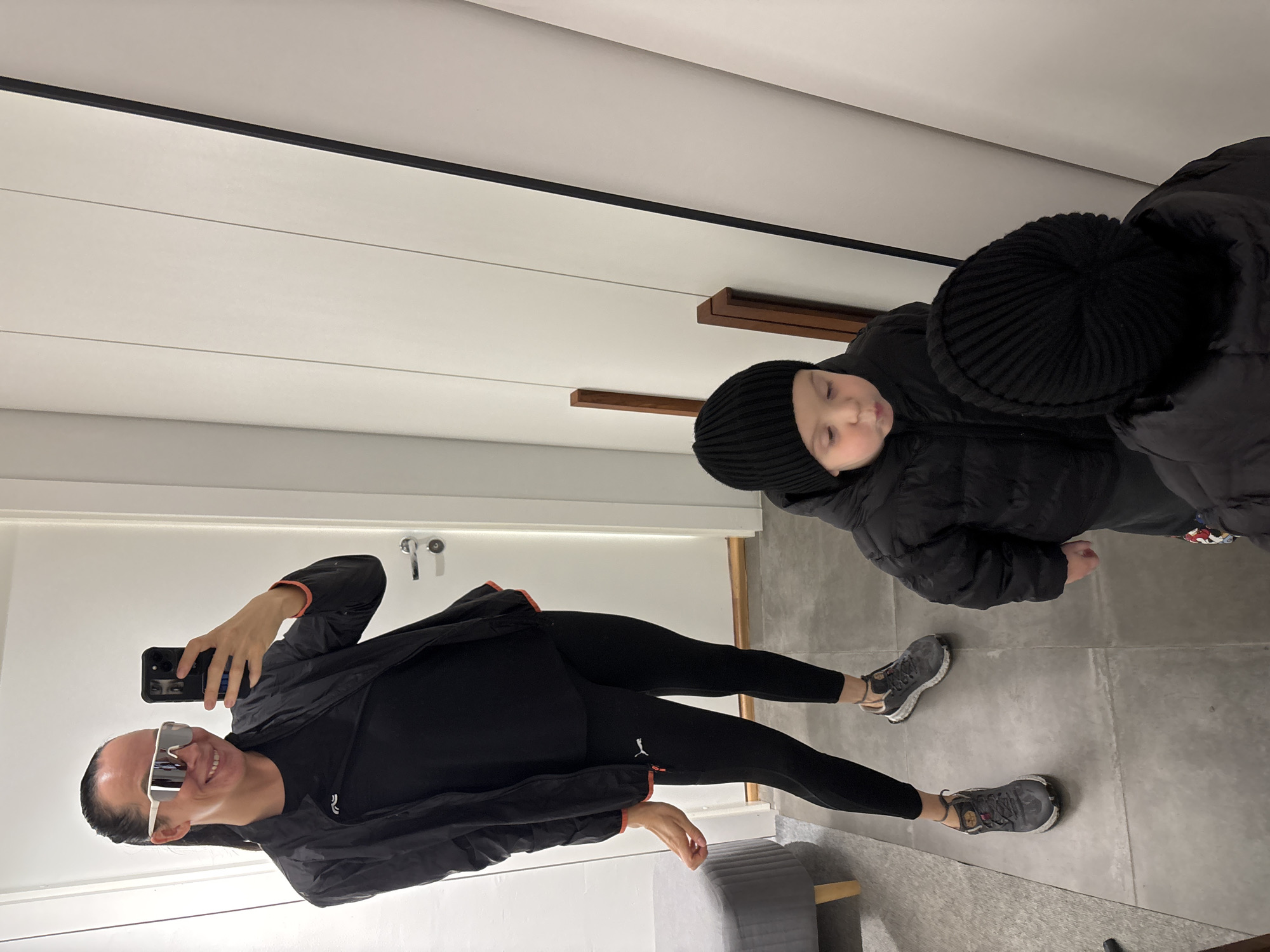 The image shows a woman taking a selfie in a dressing room. The location is likely a store fitting room. The woman is wearing sunglasses, a black jacket, black leggings, and running shoes. Beside her, a young child is wearing a black beanie and a black puffy jacket. The child appears to be tired or resting. The dressing room has white walls and a grey floor. The store's location is implied in Chaikovsky, Russia.