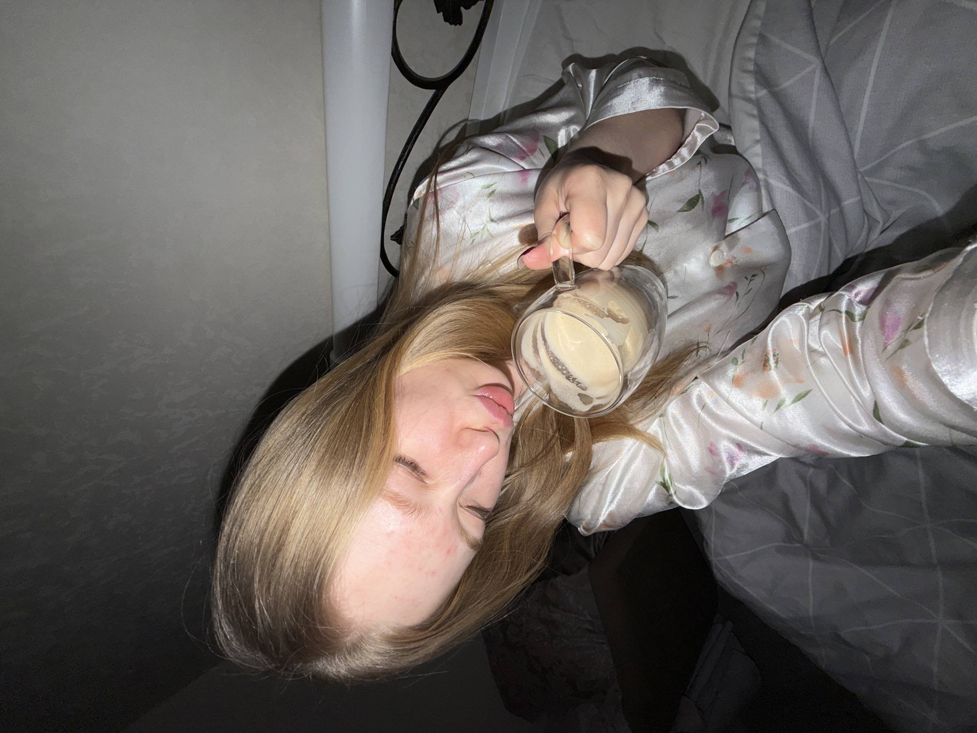 The image displays a close-up, slightly overhead shot of a young woman lying in bed, with the photo oriented 90 degrees clockwise. She has long, straight, blonde hair and appears to have her eyes closed or partially closed, conveying a sleepy or relaxed demeanor. Her face shows some minor redness or blemishes.
She is wearing light-colored, possibly white or pale pink, satin or silk-like pajamas adorned with a delicate floral pattern in pink and green. With her right hand, she holds a clear glass mug filled with a frothy, light brown beverage, likely coffee or a latte, positioned near her face.
The woman is partially covered by a grey duvet with a subtle geometric pattern. In the background, a plain light grey wall is visible, along with some dark, ornate metalwork, possibly part of a bed frame or headboard. A white vertical fixture, potentially a pipe or cable, can be seen to the left. The overall lighting is dim, suggesting an indoor setting during early morning, late evening, or under low artificial light. No visible text is present in the image. The scene evokes a feeling of calm and a leisurely start to the day.