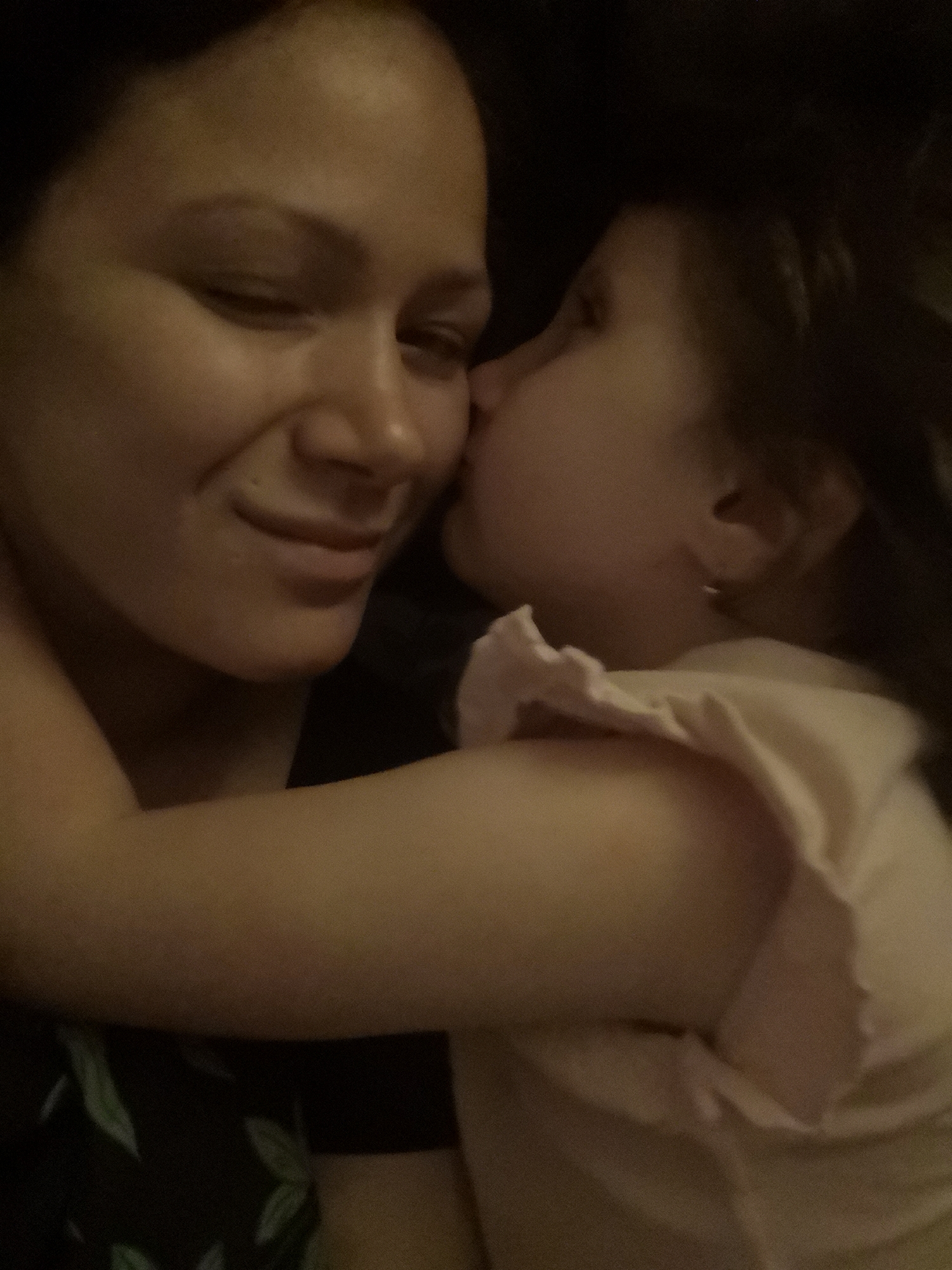 The image shows an intimate moment between two people. The primary subject is a woman whose face is visible in profile, looking to her left with her eyes closed and a gentle smile. Her skin appears smooth and illuminated. To her right, a child is shown with their face turned towards the woman, kissing her cheek. The child is wearing a light-colored garment with ruffled details on the shoulder. The background is dark, suggesting a low-light environment, possibly indoors and at night.
The activity captured is a tender interaction, a kiss given by the child to the woman, highlighting a loving relationship. The emotional tone is clearly affectionate and warm. There are no discernible objects or textual elements in the image. The overall impression is one of closeness and familial love. The lighting is soft, casting a warm glow on the subjects' faces and emphasizing their connection. The dark background serves to focus attention on the individuals and their embrace.