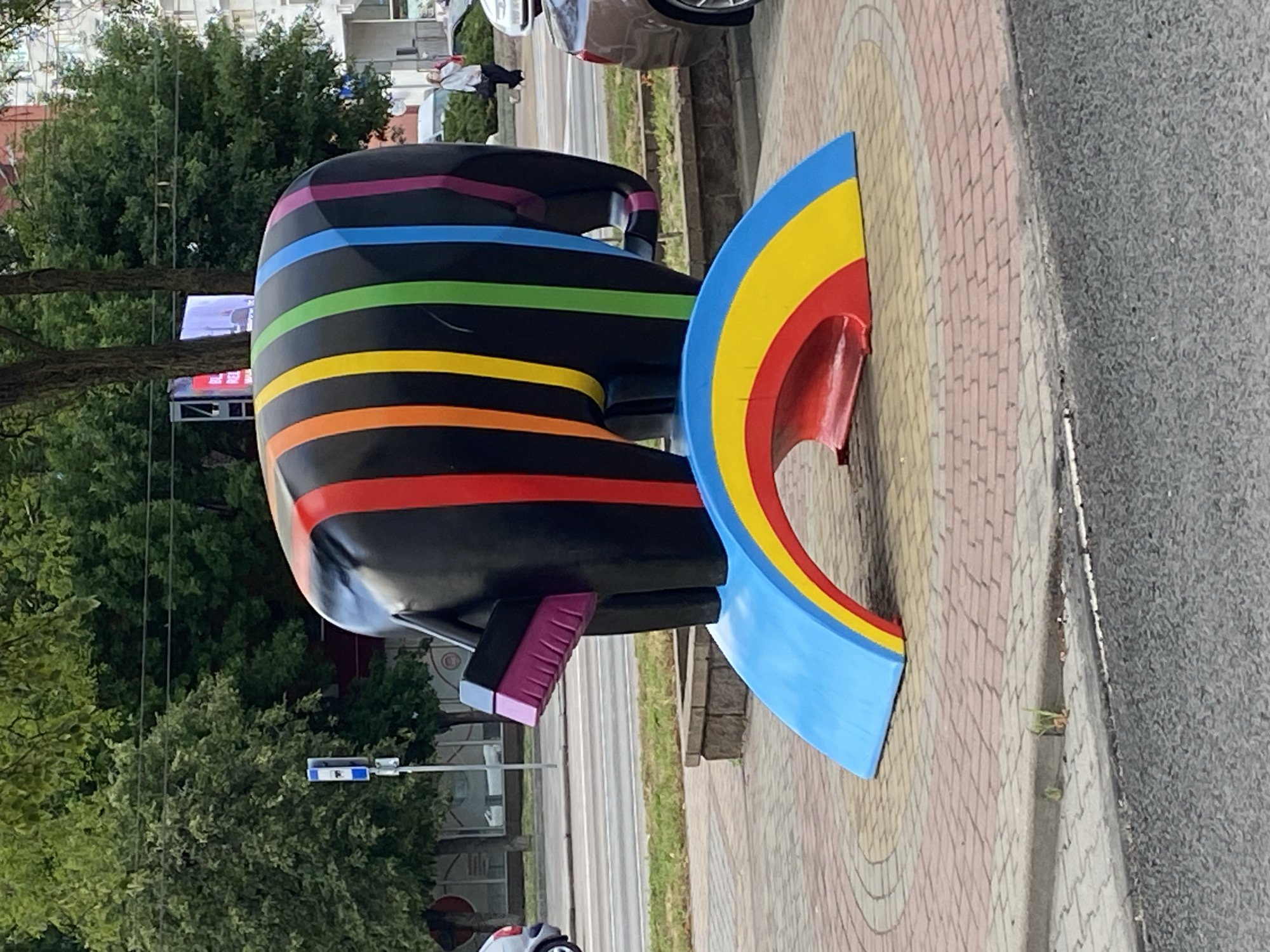 A large, black, abstract sculpture resembling an elephant with rainbow-colored stripes is displayed outdoors on a patterned brick circular base. The elephant appears to be lifting a broom with a purple brush and pink handle with its trunk. A segment of a rainbow, painted in red, yellow, and blue, is positioned beneath the elephant. The sculpture is located on a sidewalk in front of a street, with trees and buildings visible in the background. A woman is walking in the distance on the sidewalk. The location is the city of Brest, Belarus.