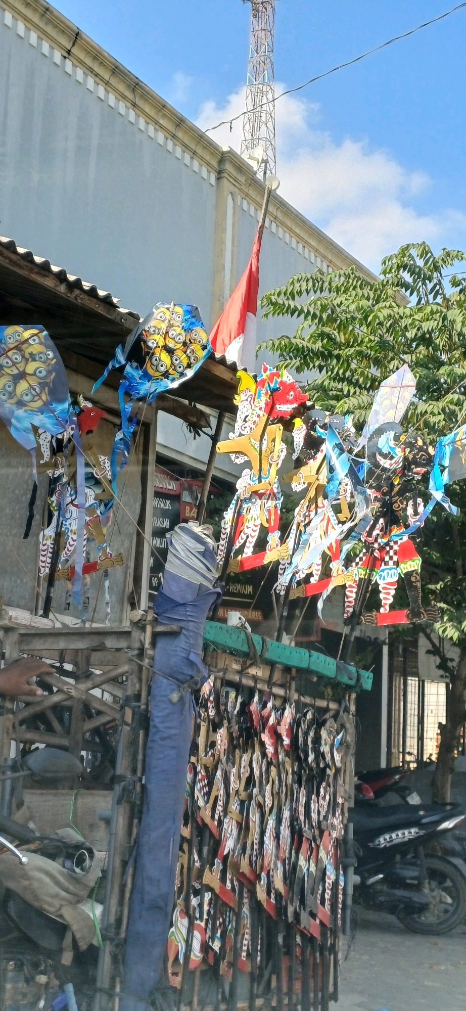 In Pucakwangi, Indonesia, a storefront displays an array of traditional Indonesian wayang kulit (shadow puppets) and kites for sale. The puppets, made of carved leather, are intricately decorated and hang in rows. Above them, colorful kites with cartoon characters, including minions, are displayed. A red and white Indonesian flag is visible in the background, attached to a tall communication tower. The scene appears to be outdoors, with a building and trees providing a backdrop. A motorcycle is parked in front of the store, suggesting it's a busy street or market area. The overall impression is one of local commerce and cultural heritage.